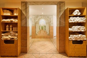 A hotel lobby with a central marble pathway, wooden shelving with towels and toiletries, and a classical archway leading to a distant view.