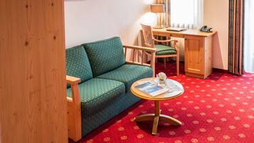 Une pièce avec un canapé vert, un bureau en bois, une chaise, une petite table ronde sur laquelle se trouve un magazine, et un tapis rouge à motifs.