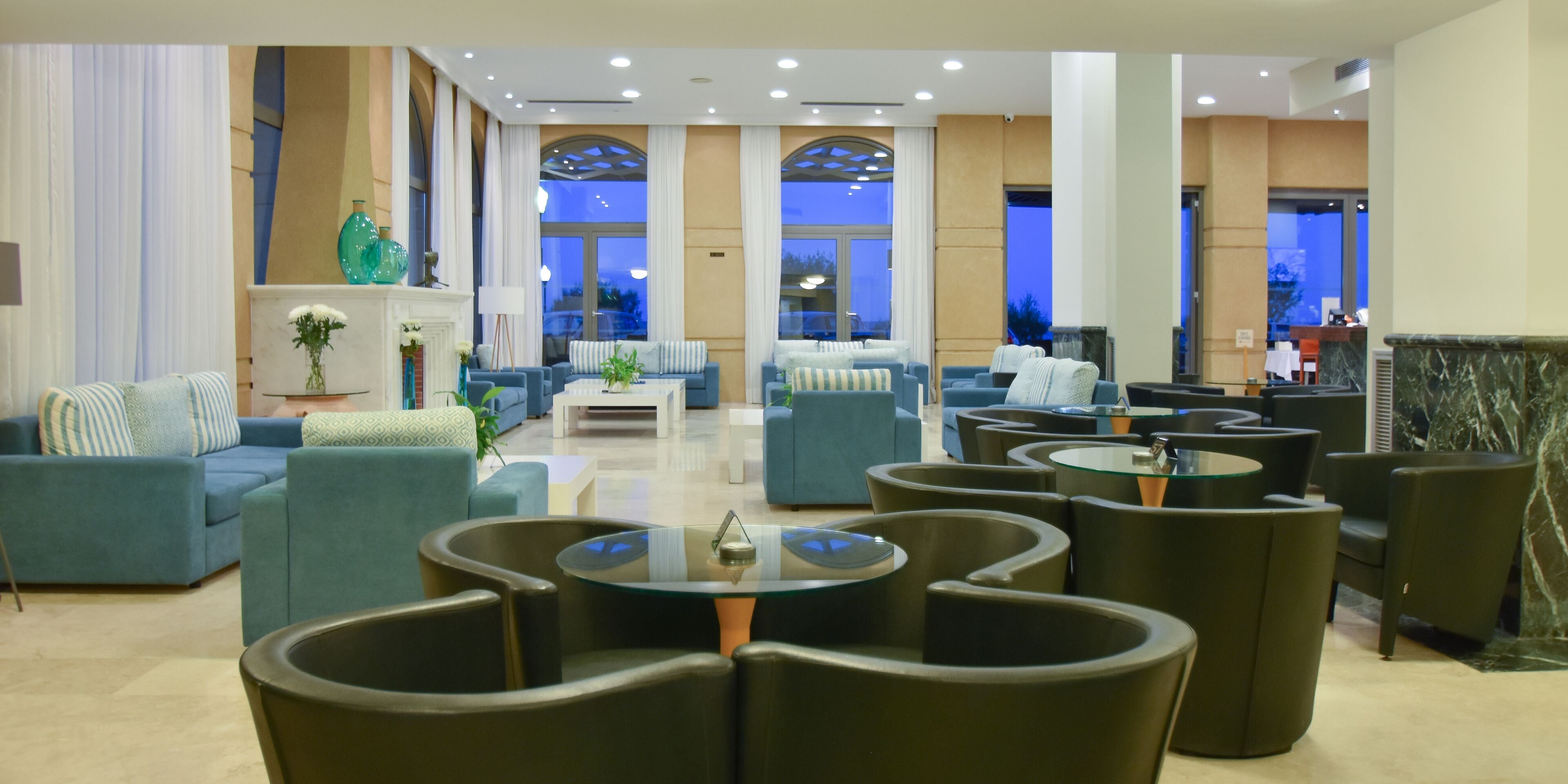 A spacious lobby with multiple seating areas, including sofas and round tables, and large windows offering views of the outside.