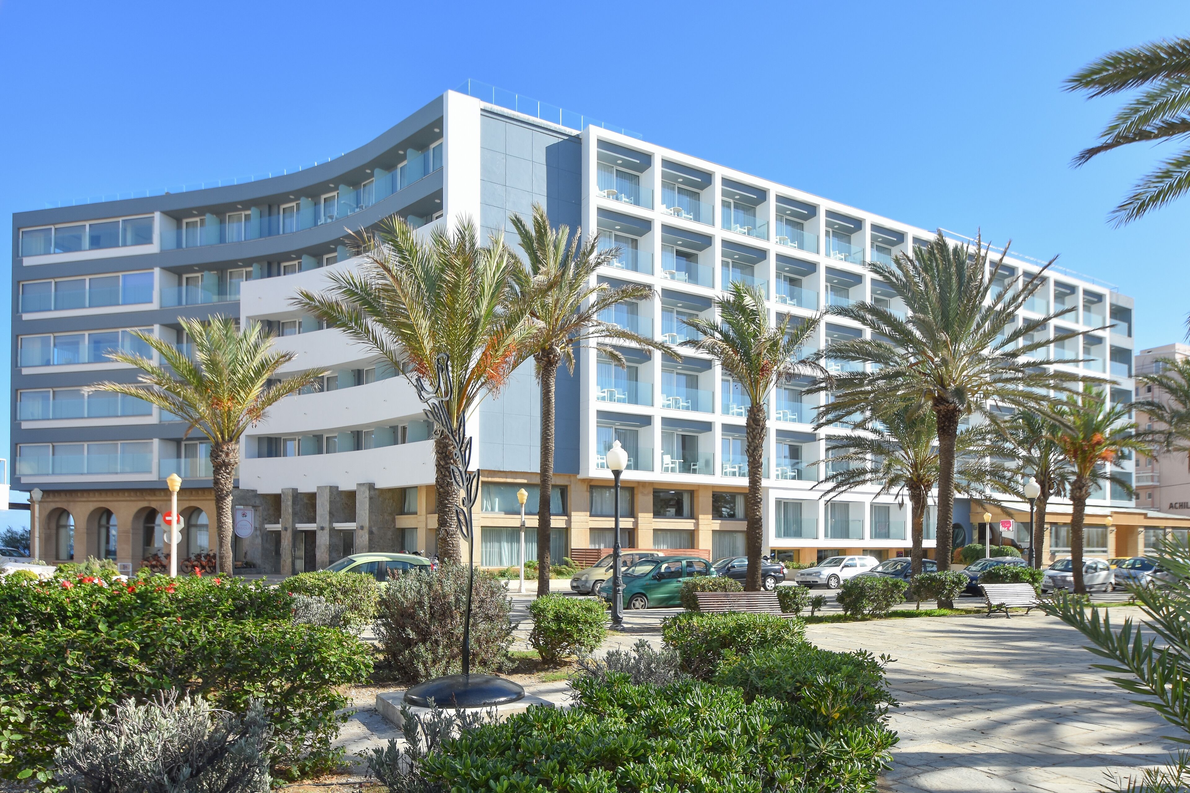 A modern hotel with a curved facade, multiple balconies, and a landscaped courtyard with palm trees and shrubs.