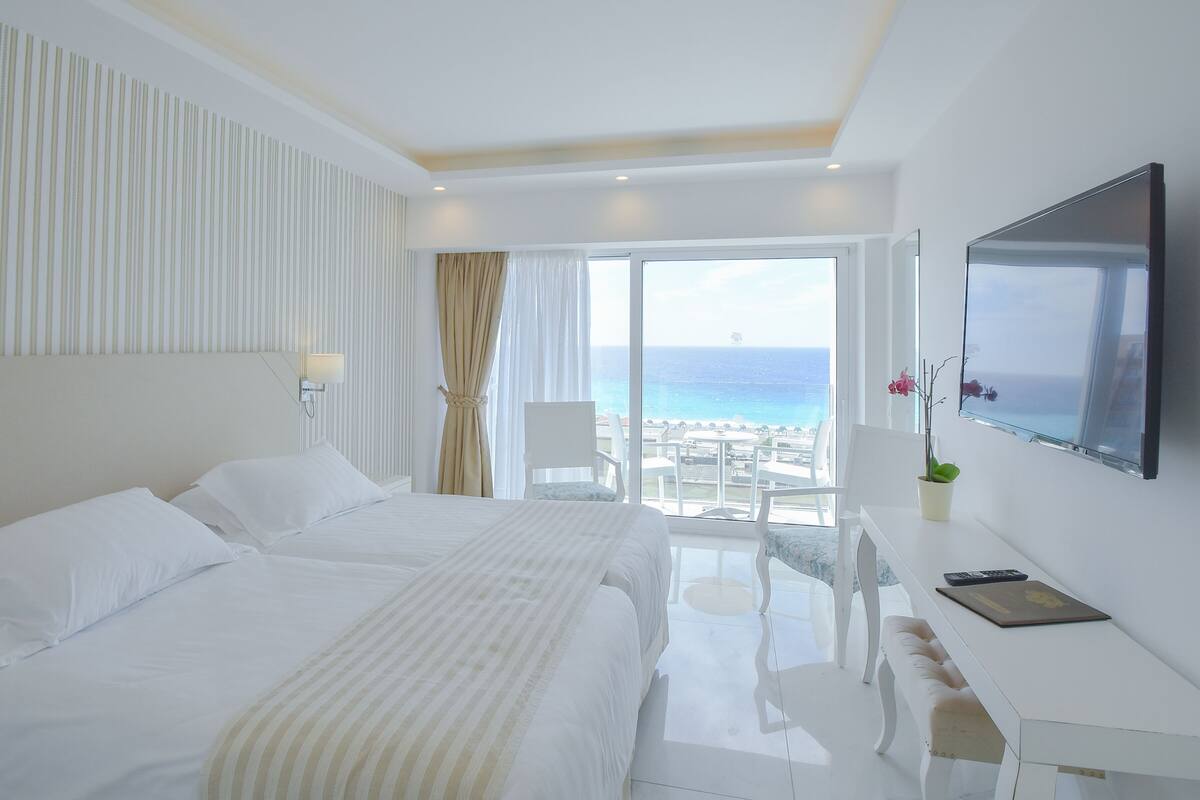 A hotel room with a large bed, a desk with a laptop, a TV, and a view of the beach.