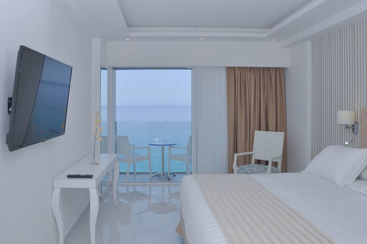 A hotel room with a bed, a desk with a TV, a chair, a small table, and a view of the sea.
