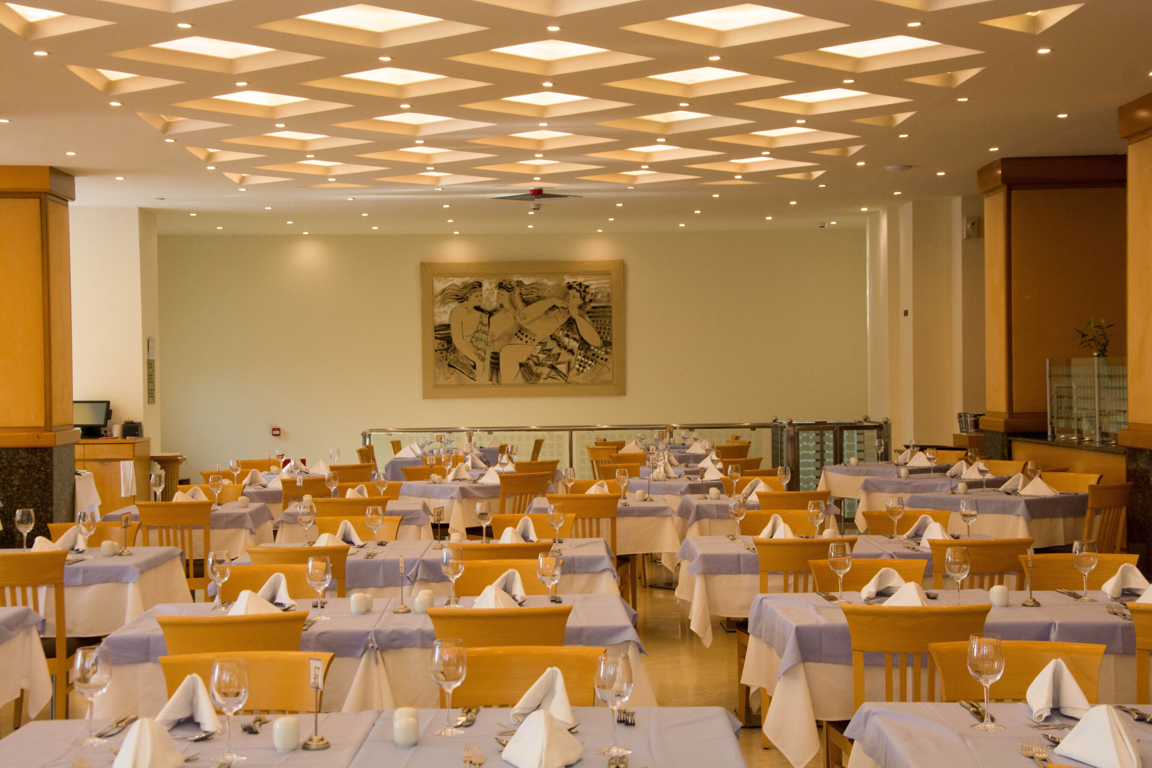 Un comedor con varias mesas dispuestas para los huéspedes, con un diseño geométrico en el techo y una gran obra de arte en la pared.