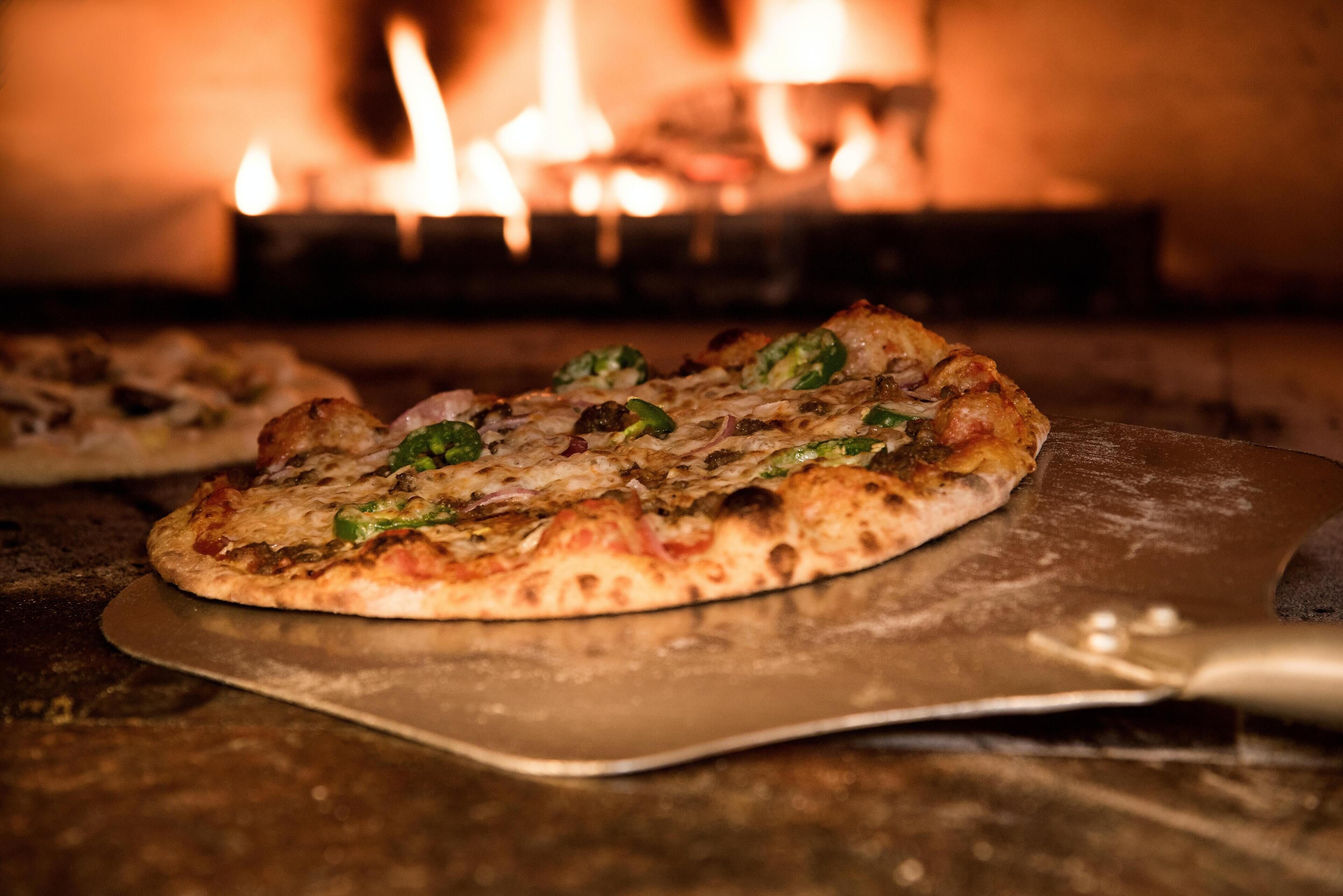 A pizza on a metal tray with a wooden table, in front of a fireplace with visible flames.