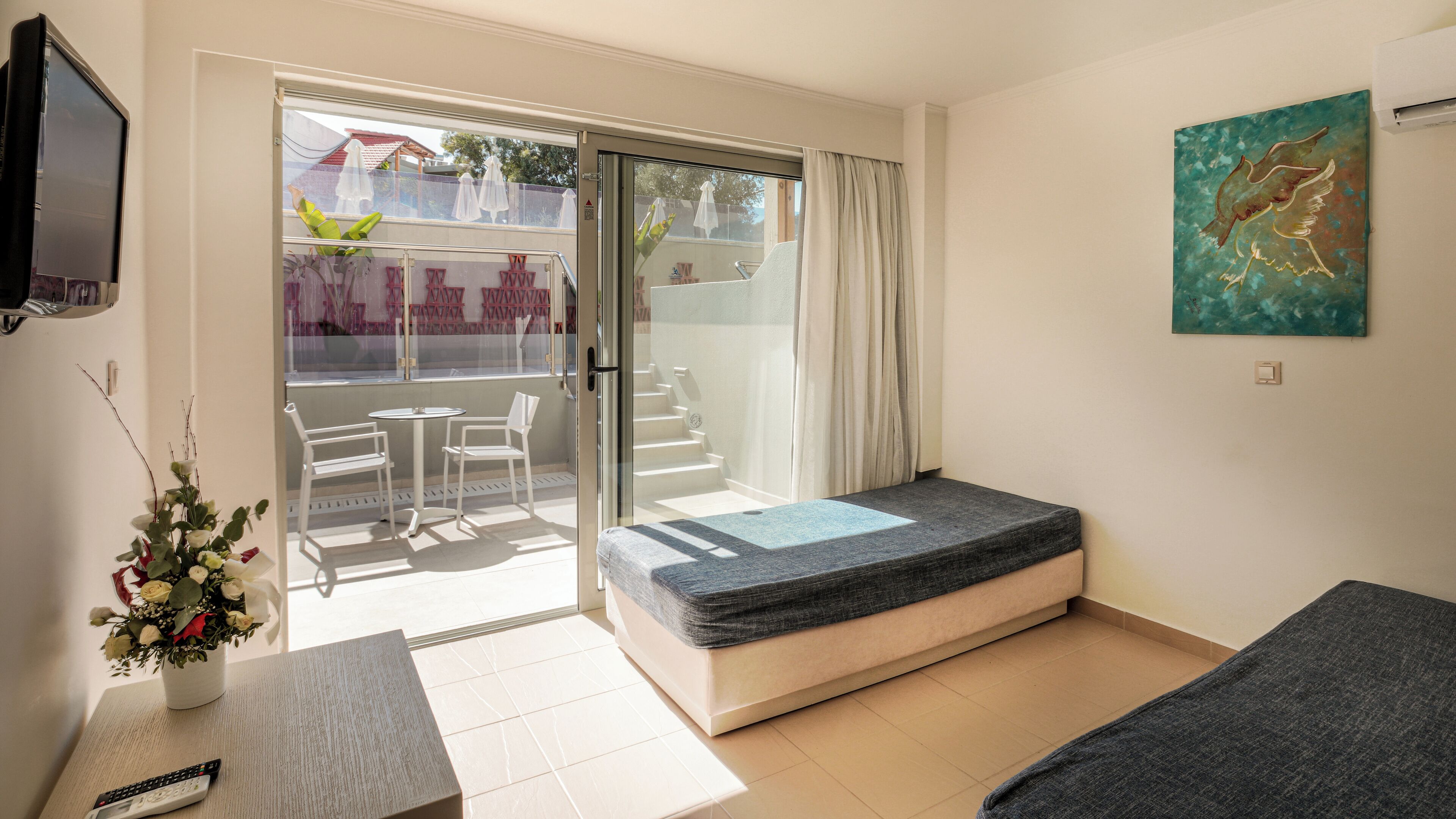 Ein kleiner, moderner Raum mit Bett, Fernseher, Tisch mit Stühlen und Blick auf einen Balkon mit Gartenmöbeln.