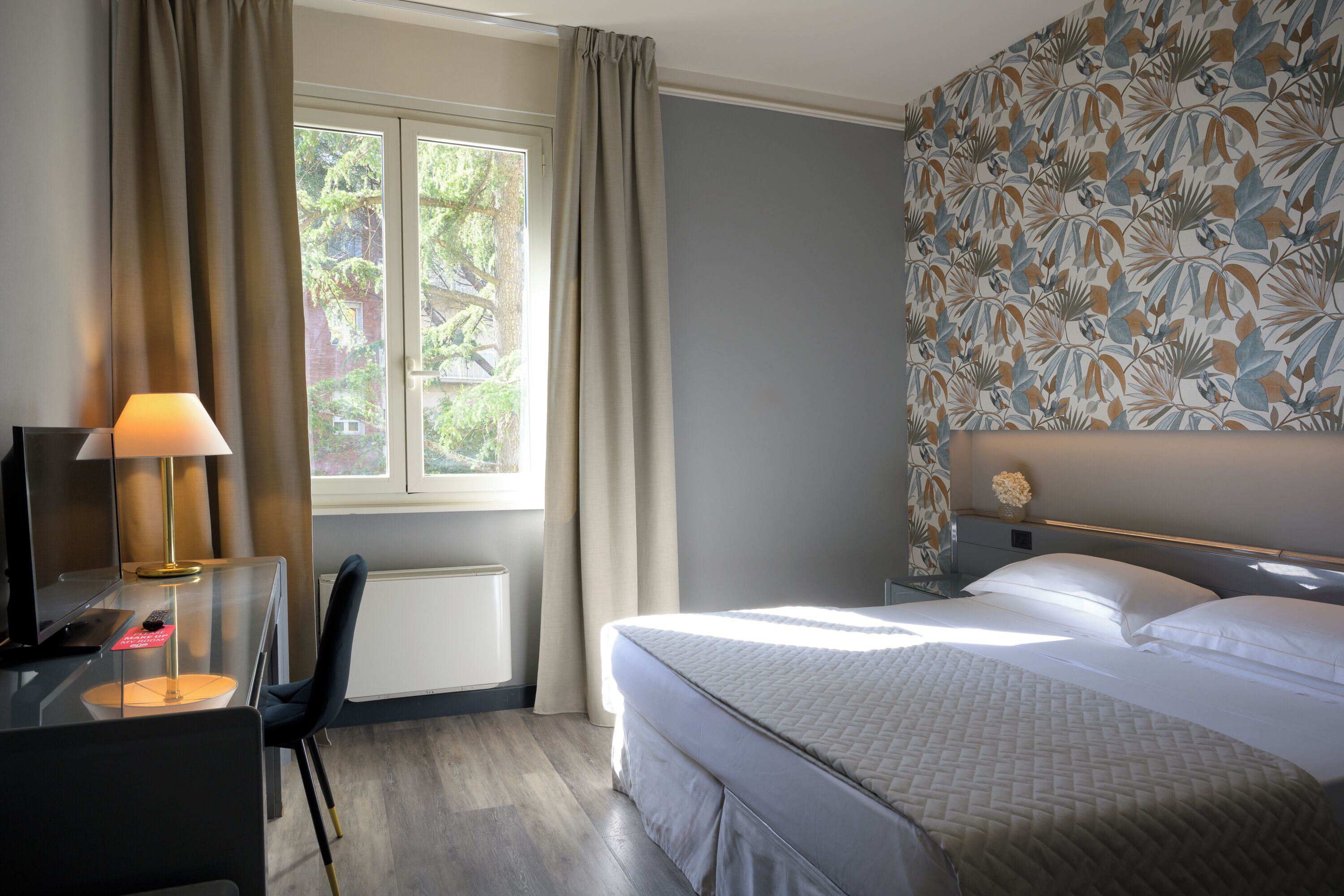A hotel room with a bed, a desk with a lamp, a chair, a window with curtains, and a wall with a leaf pattern.