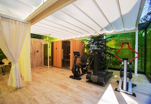 A modern gym with exercise equipment, a wooden floor, and a glass wall offering a view of greenery. - Grand Hotel Panoramic (Montecatini Terme)