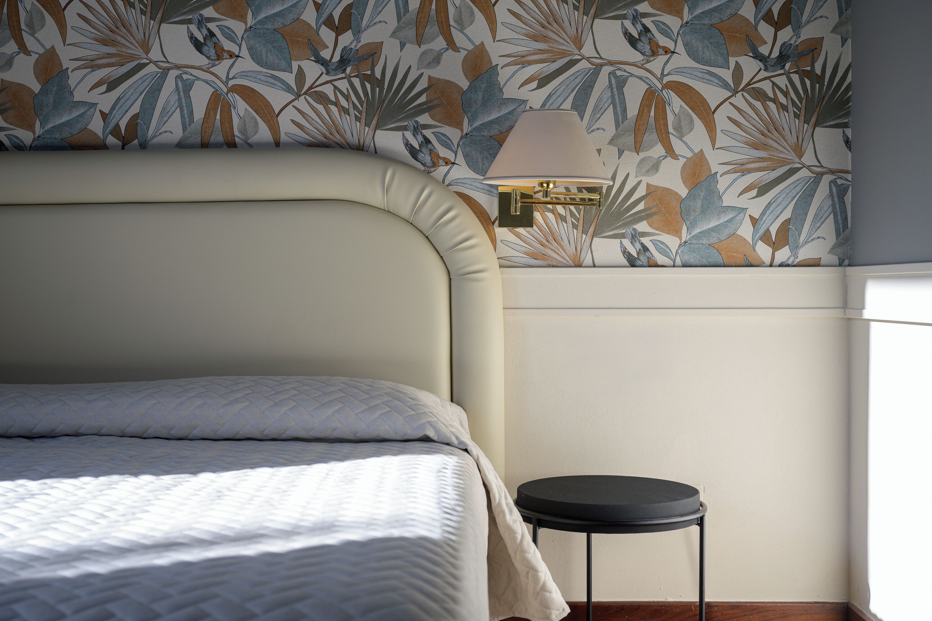 A neatly made bed with a headboard, a small round stool, and a wall with a floral wallpaper pattern.