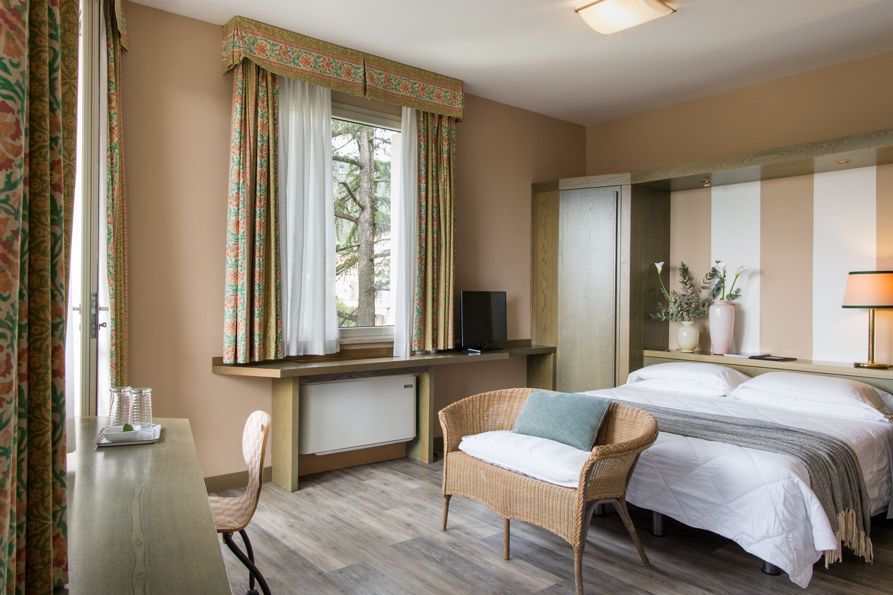 A bedroom with a bed, a chair, a desk with a computer, a window with curtains, and a mirror.
