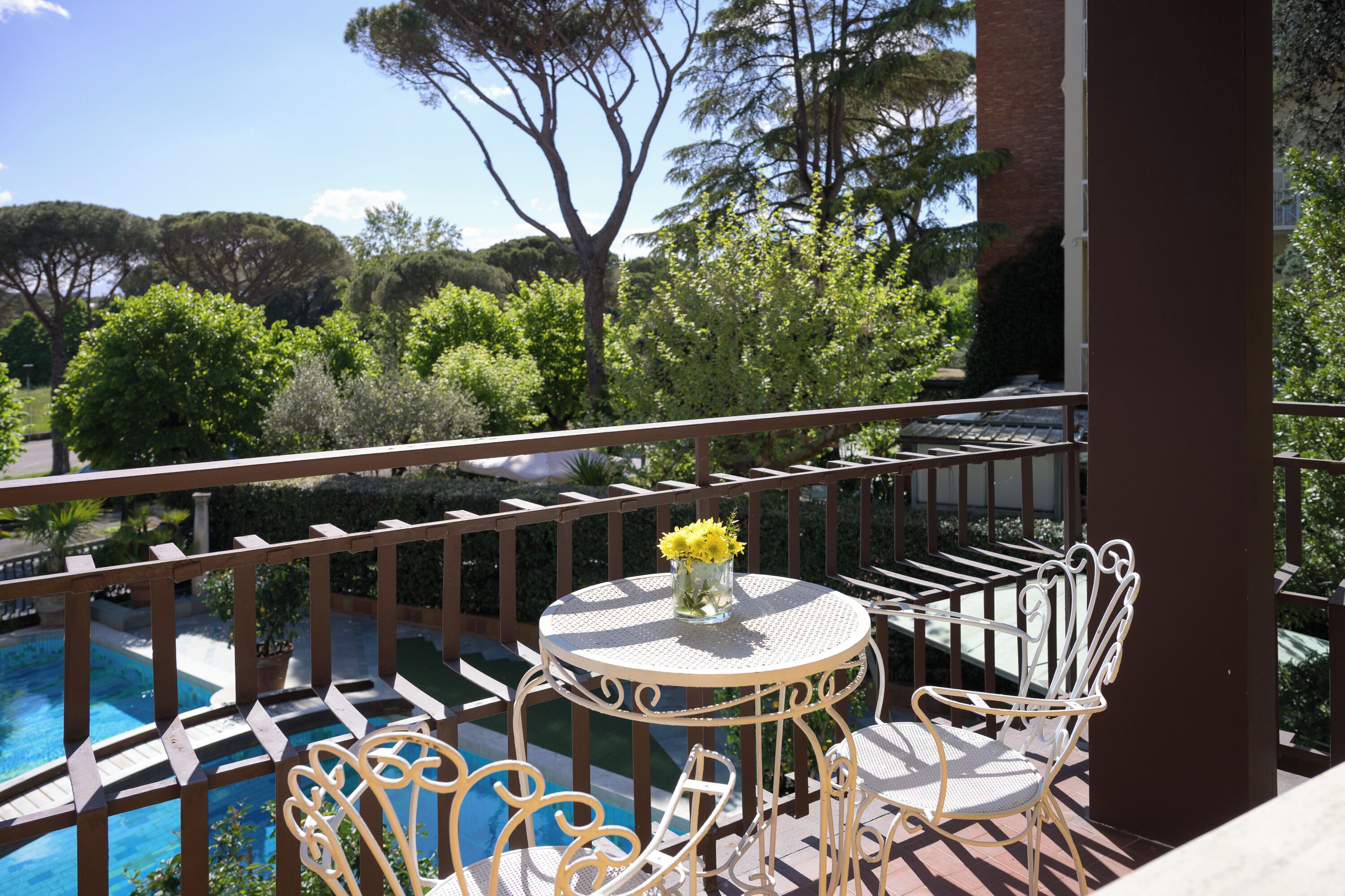 A balcony with a round table and two white chairs, overlooking a swimming pool and lush greenery.