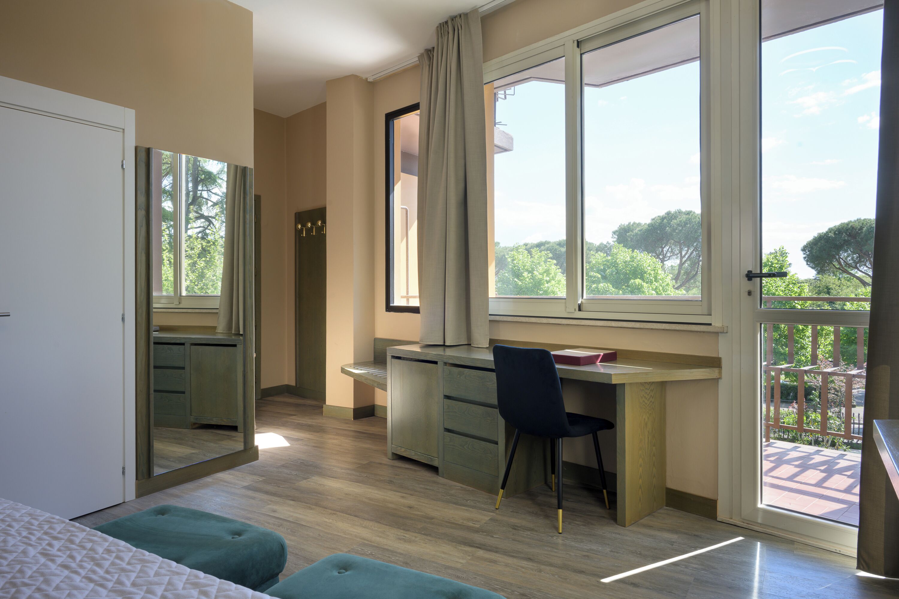 A bedroom with a bed, a desk, and a chair, a large window with a view of trees, and a balcony.