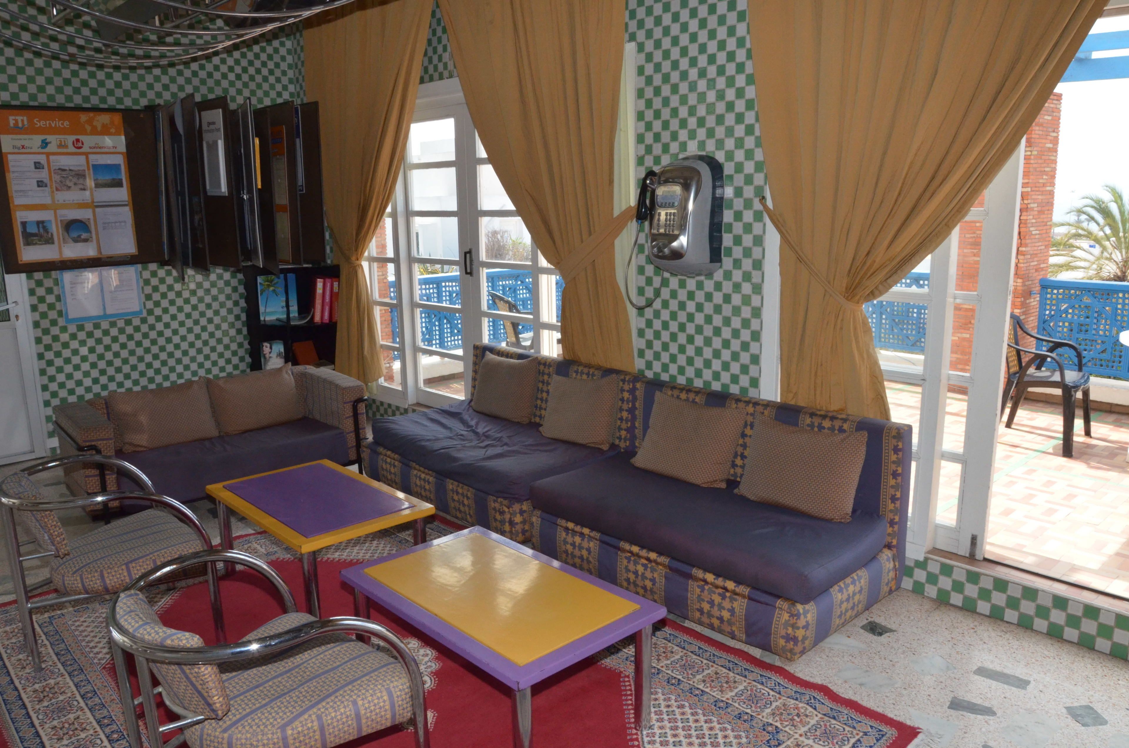 A room with a checkered floor, a sofa, two armchairs, and a small table. There is a window with curtains, a wall-mounted phone, and a bulletin board with various notices.