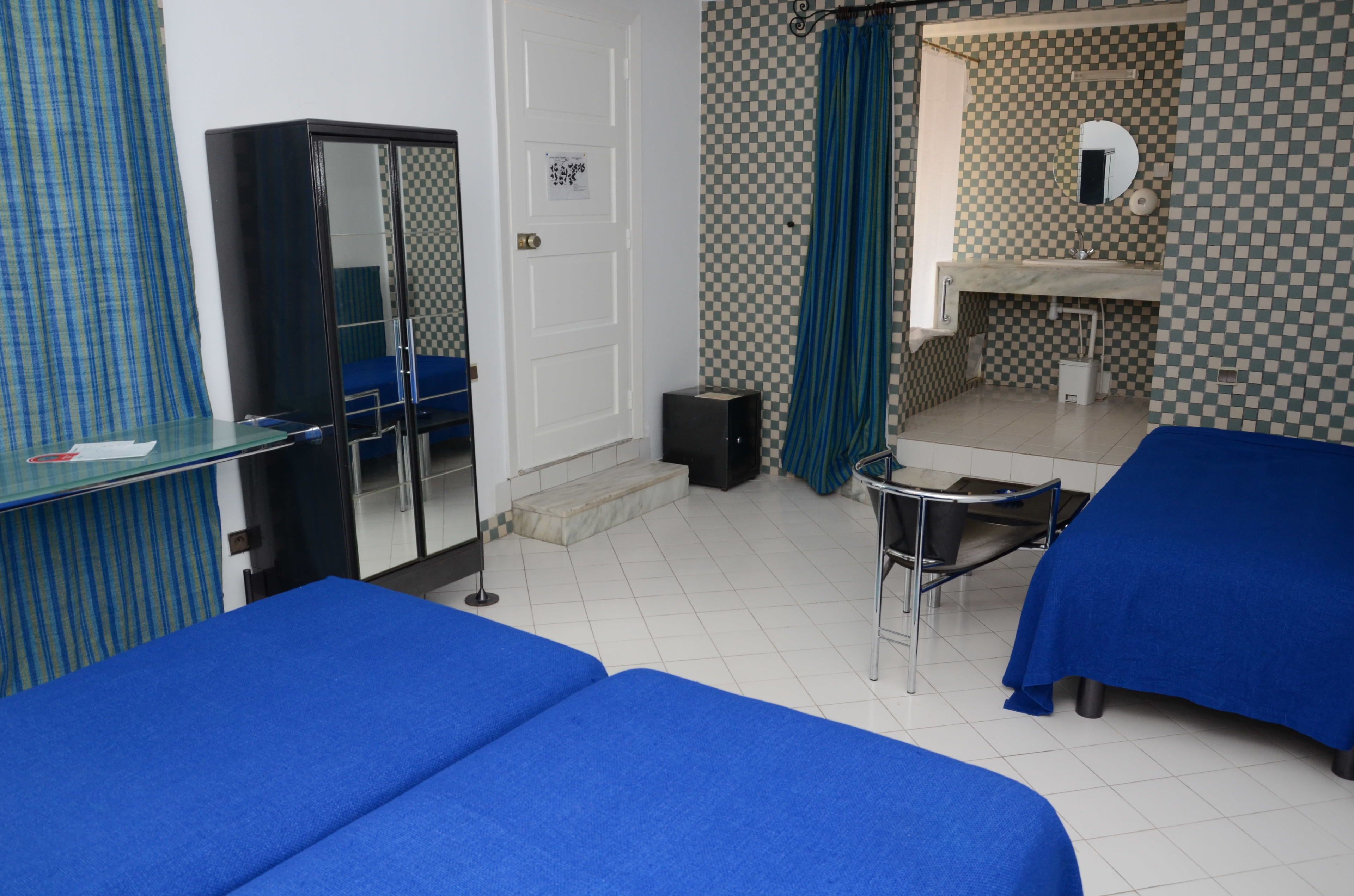 A room with two beds, a desk, a mirror, a wardrobe, and a bathroom with a checkered floor and blue curtains.