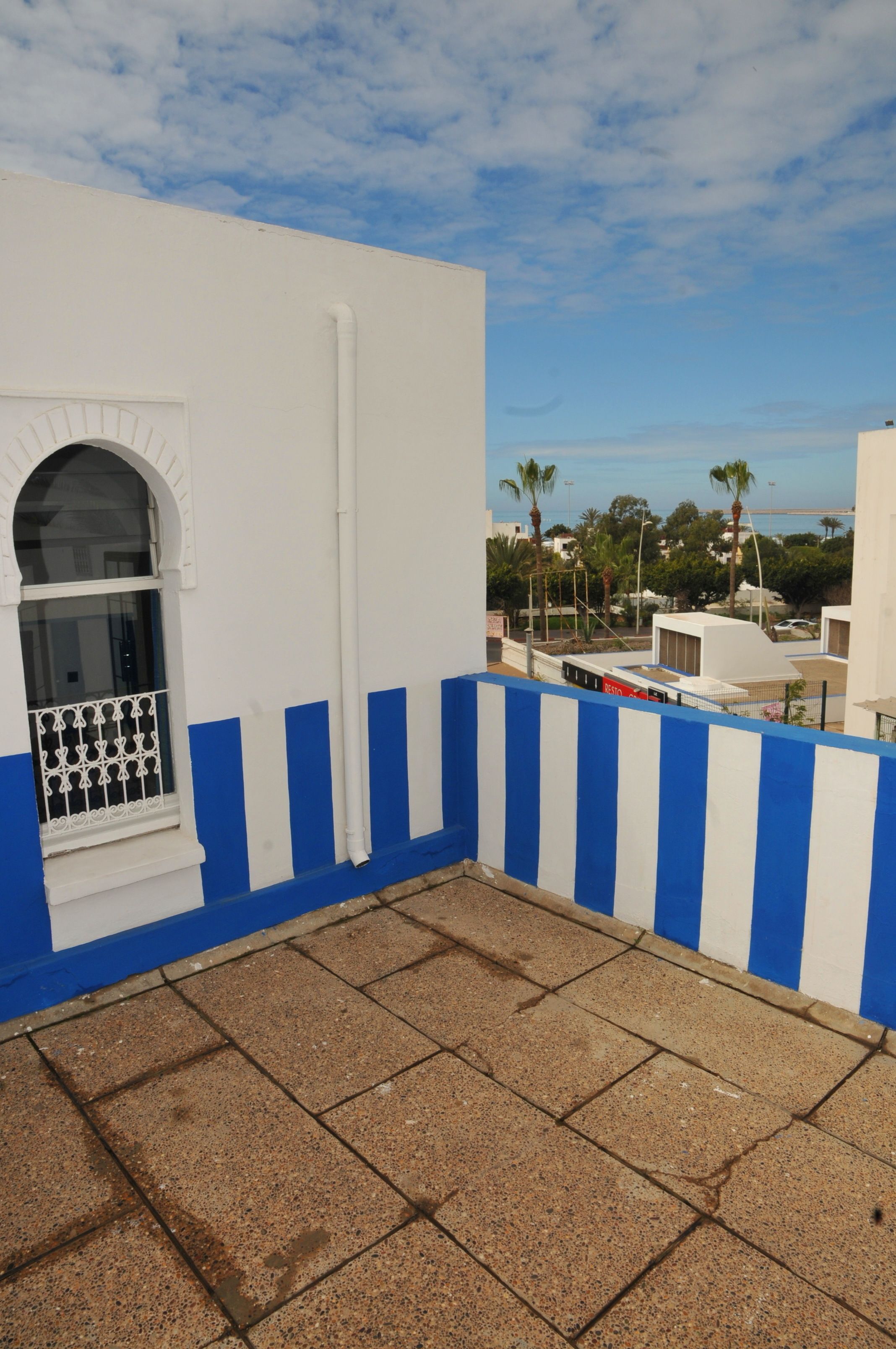 Uma varanda com piso de azulejos, uma parede branca com listras azuis e uma janela com grade decorativa.
