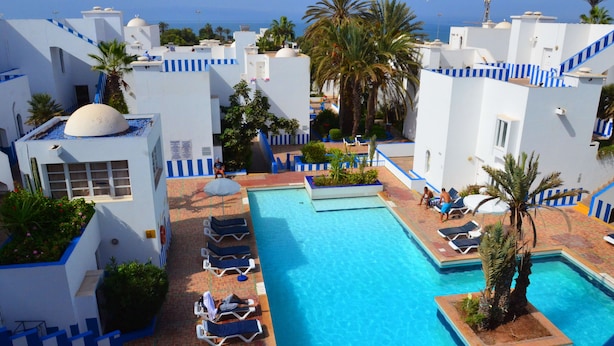 A pool area with lounge chairs and palm trees, surrounded by white buildings with blue accents. - Tagadirt Appart-Hotel (Agadir)