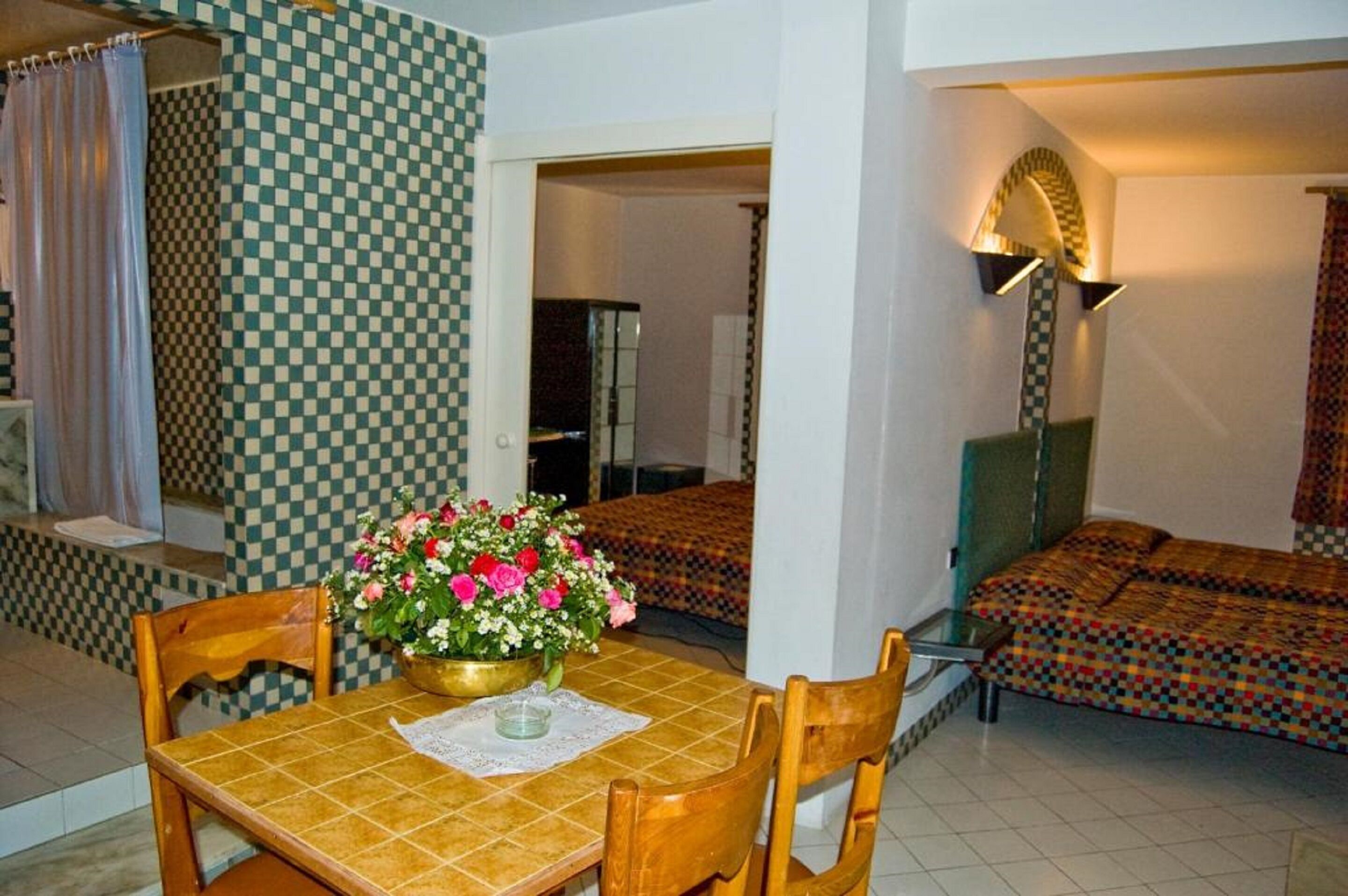Una habitación con pared a cuadros, una mesa de comedor con sillas, un florero, una cama con ropa de cama estampada y una lámpara de pie.