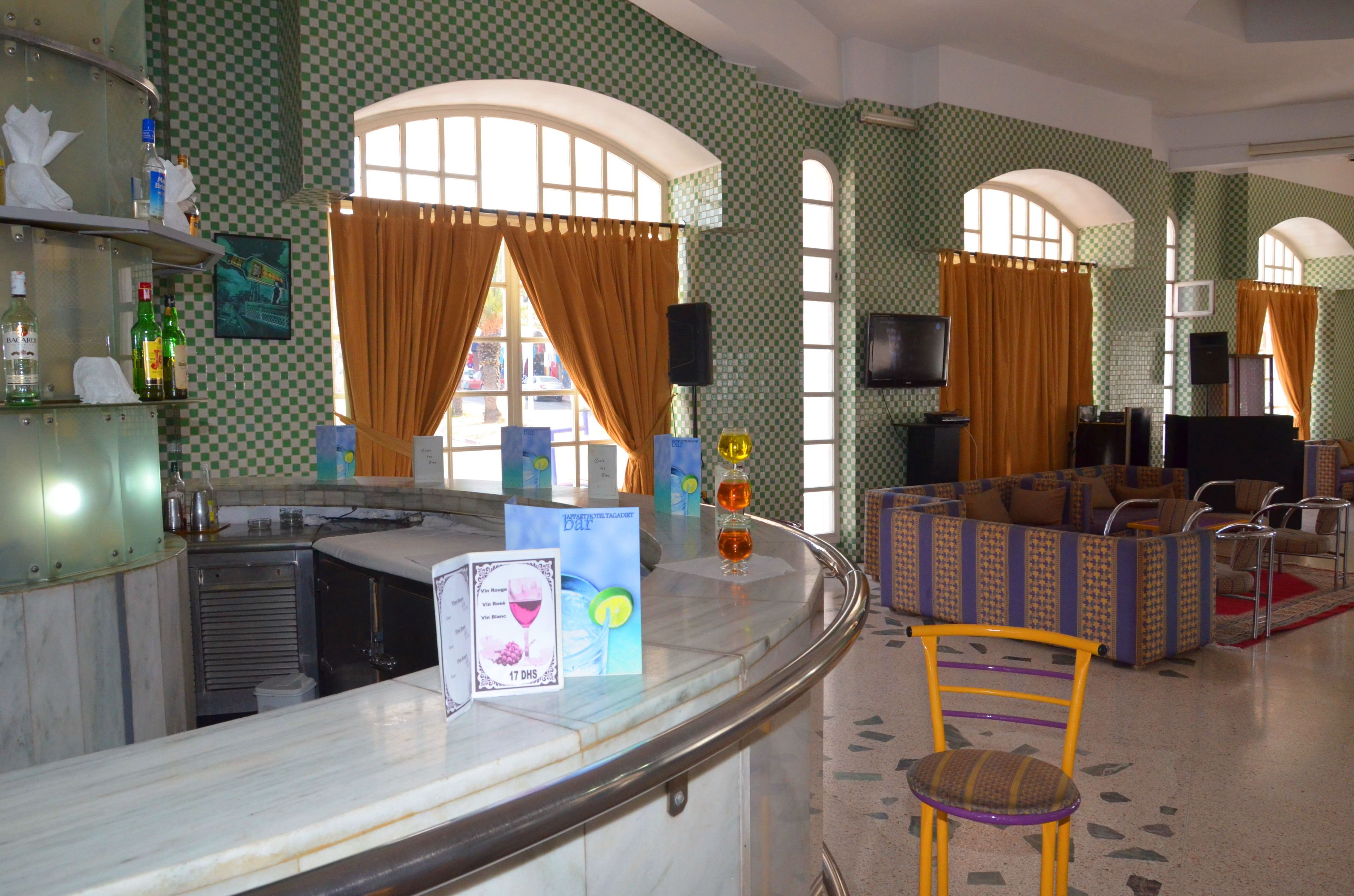 A bar area with a counter, bar stools, and a seating area with couches and chairs. There are large windows with curtains, a television, and a menu stand.