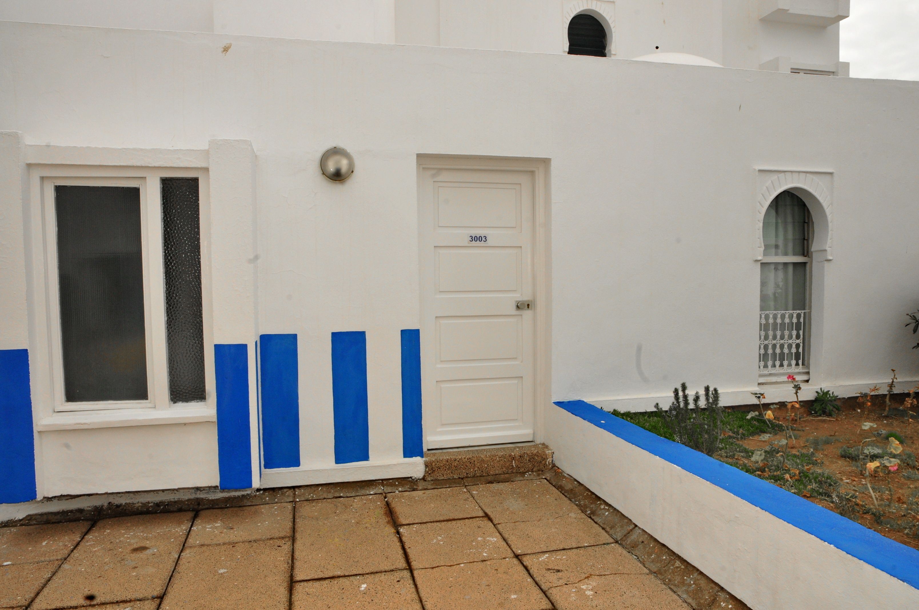 Um edifício branco com uma porta azul identificada com o número 3003, uma janela com grade de metal e uma pequena área de jardim com plantas e canteiro de flores.
