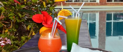 Une scène au bord de la piscine, avec deux boissons sur un plateau, l’une orange et l’autre verte, entourées de fleurs rouges et de verdure.