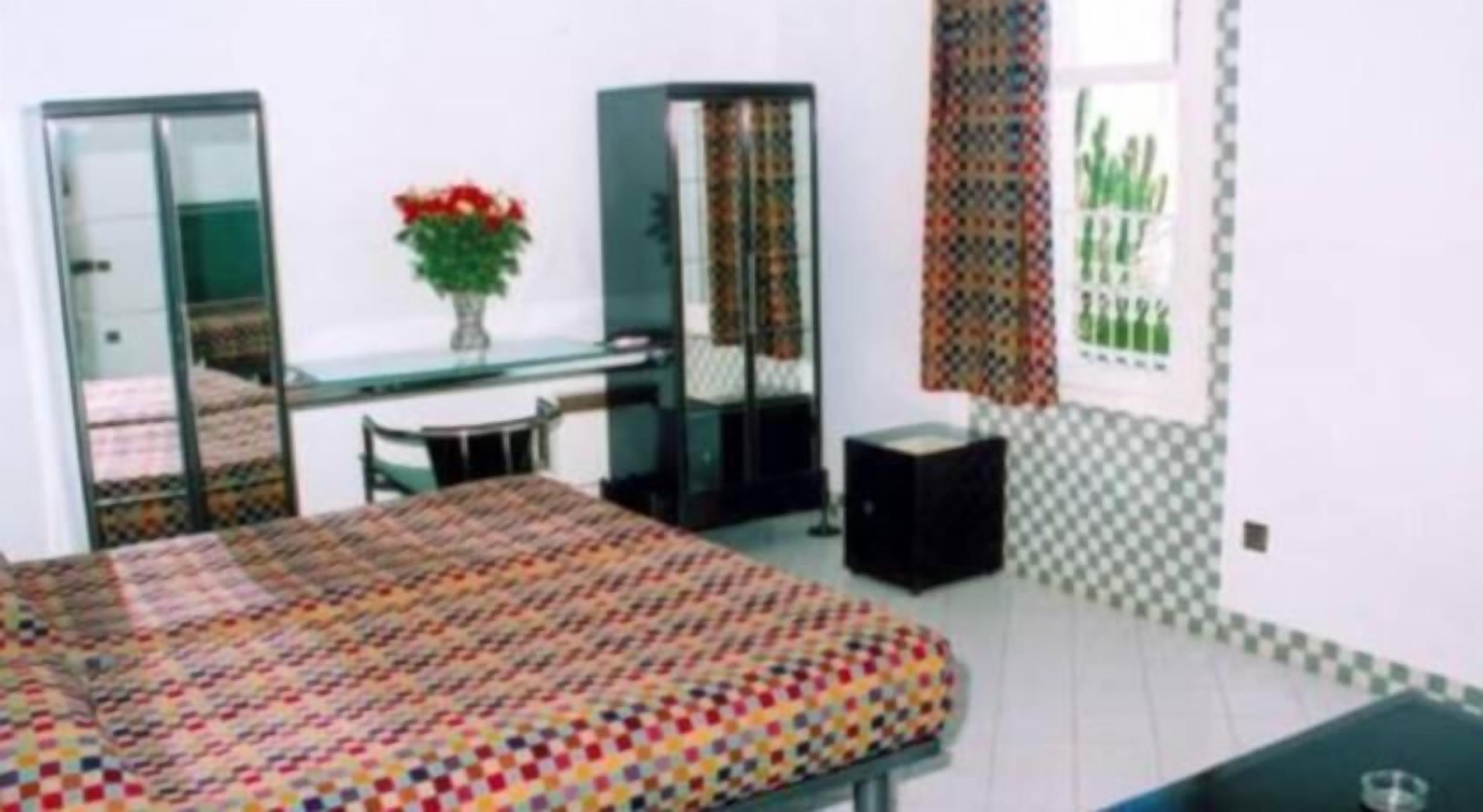 A room with a bed, a desk, a chair, a small table, a potted plant, and a window with patterned curtains.