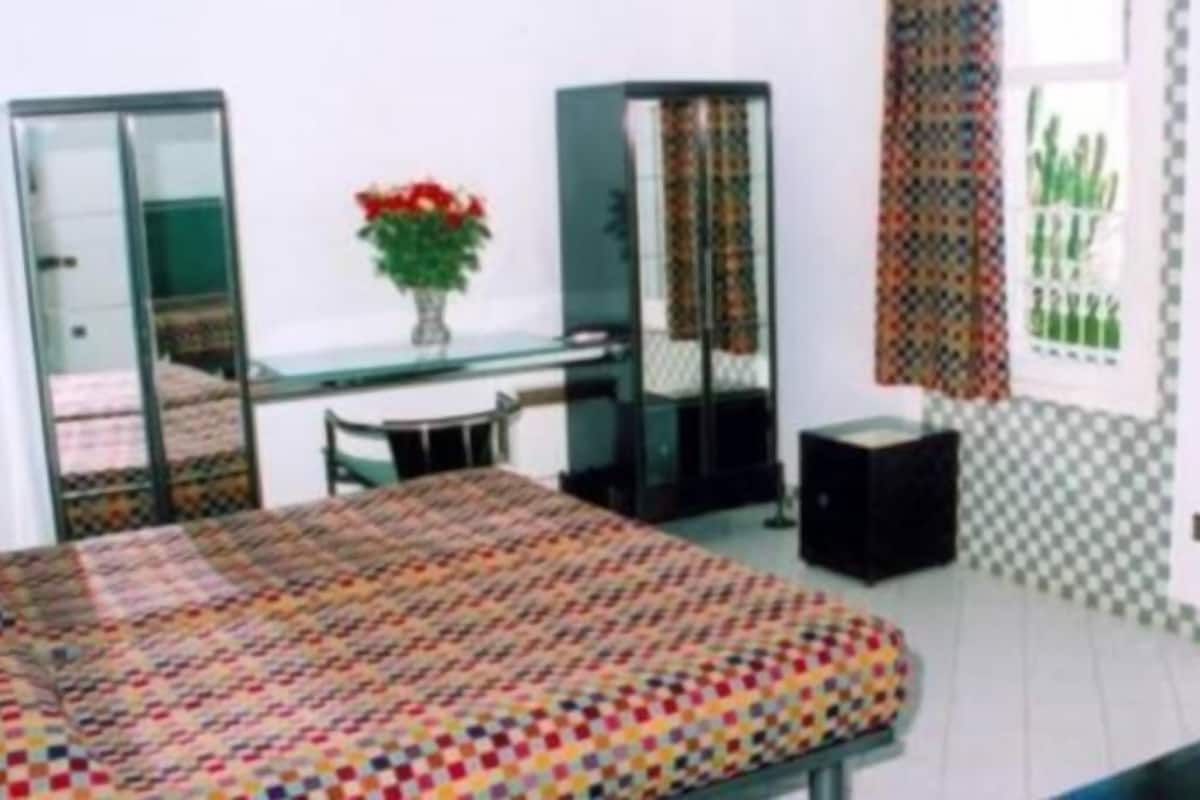 A room with a bed, a desk, a chair, a small table, a potted plant, and a window with patterned curtains.