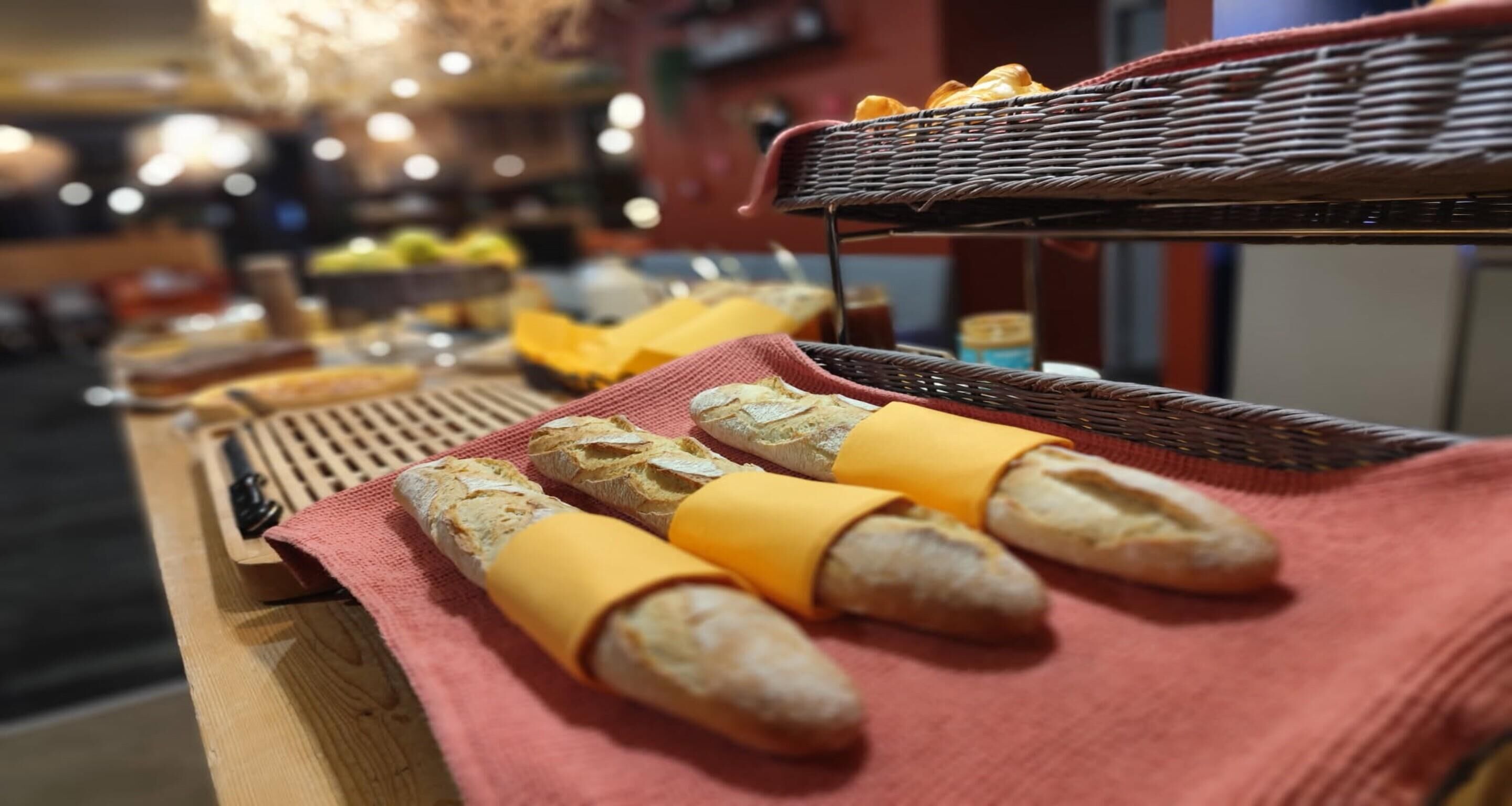 A wooden table with baguettes wrapped in yellow cheese, a wicker basket, and a red cloth.