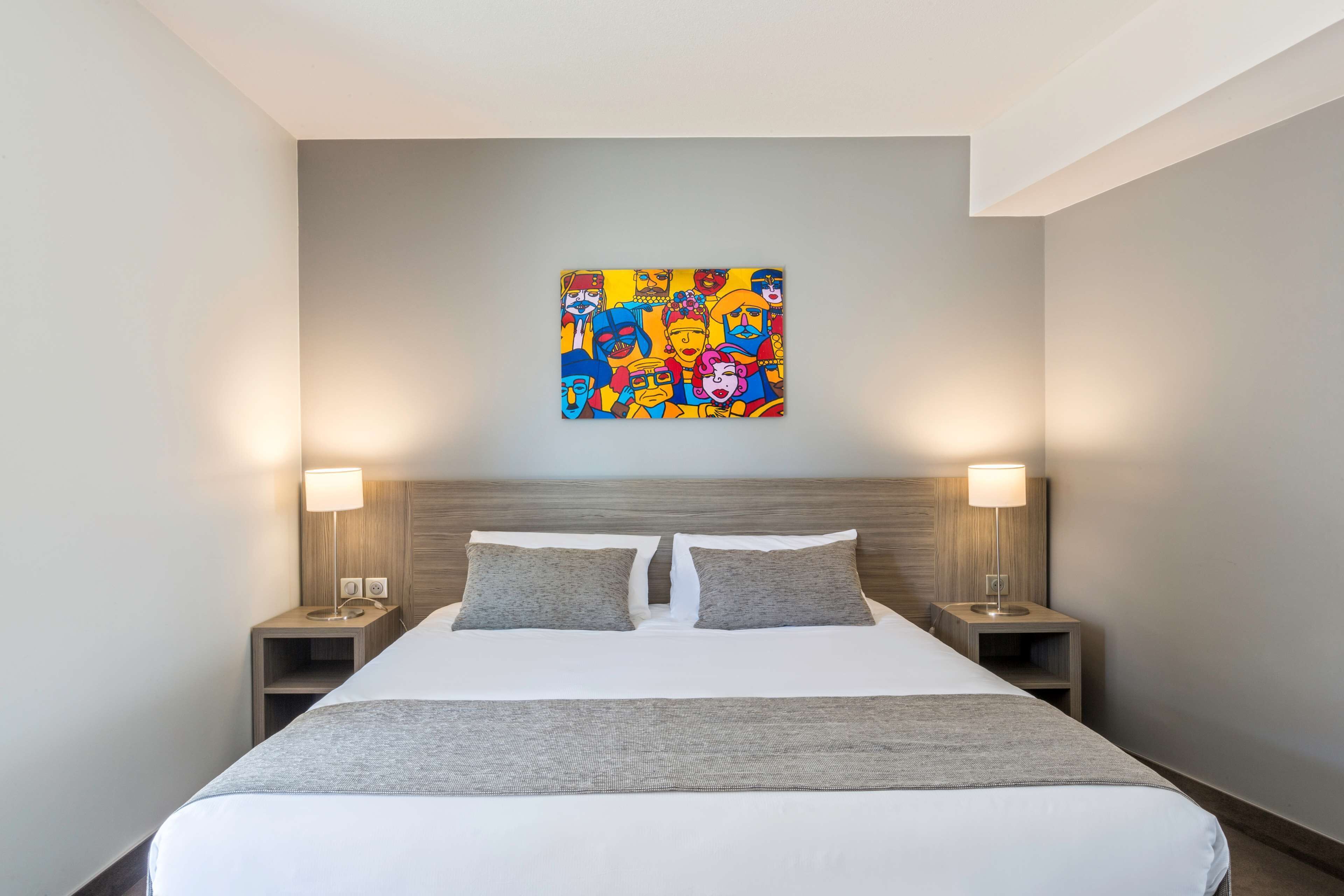 Une chambre d’hôtel avec un grand lit, deux tables de chevet avec des lampes et une peinture abstraite aux couleurs vives accrochée au mur.