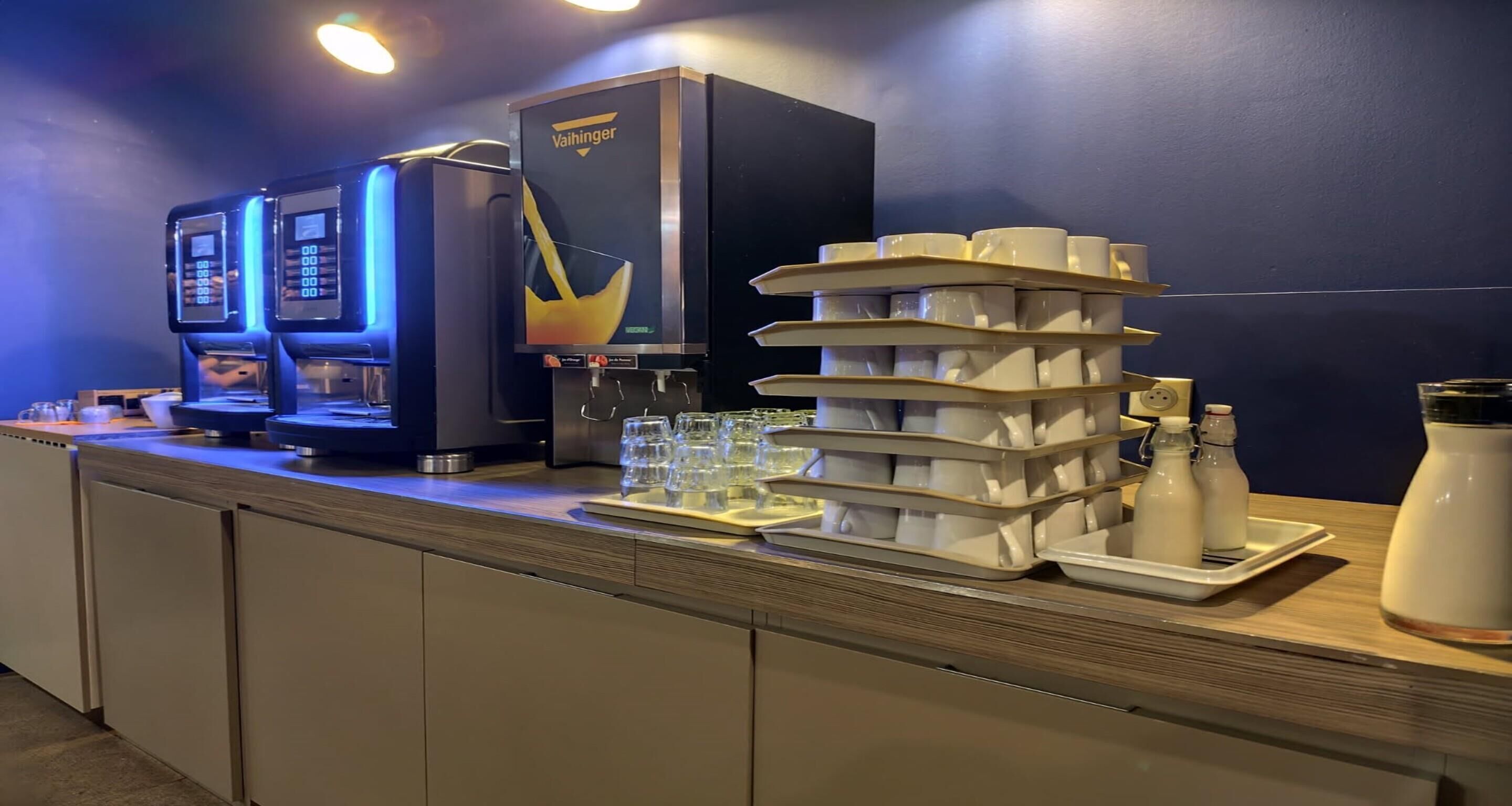 A coffee station with a Vaihinger coffee machine, a stack of disposable cups, and a tray with glasses.