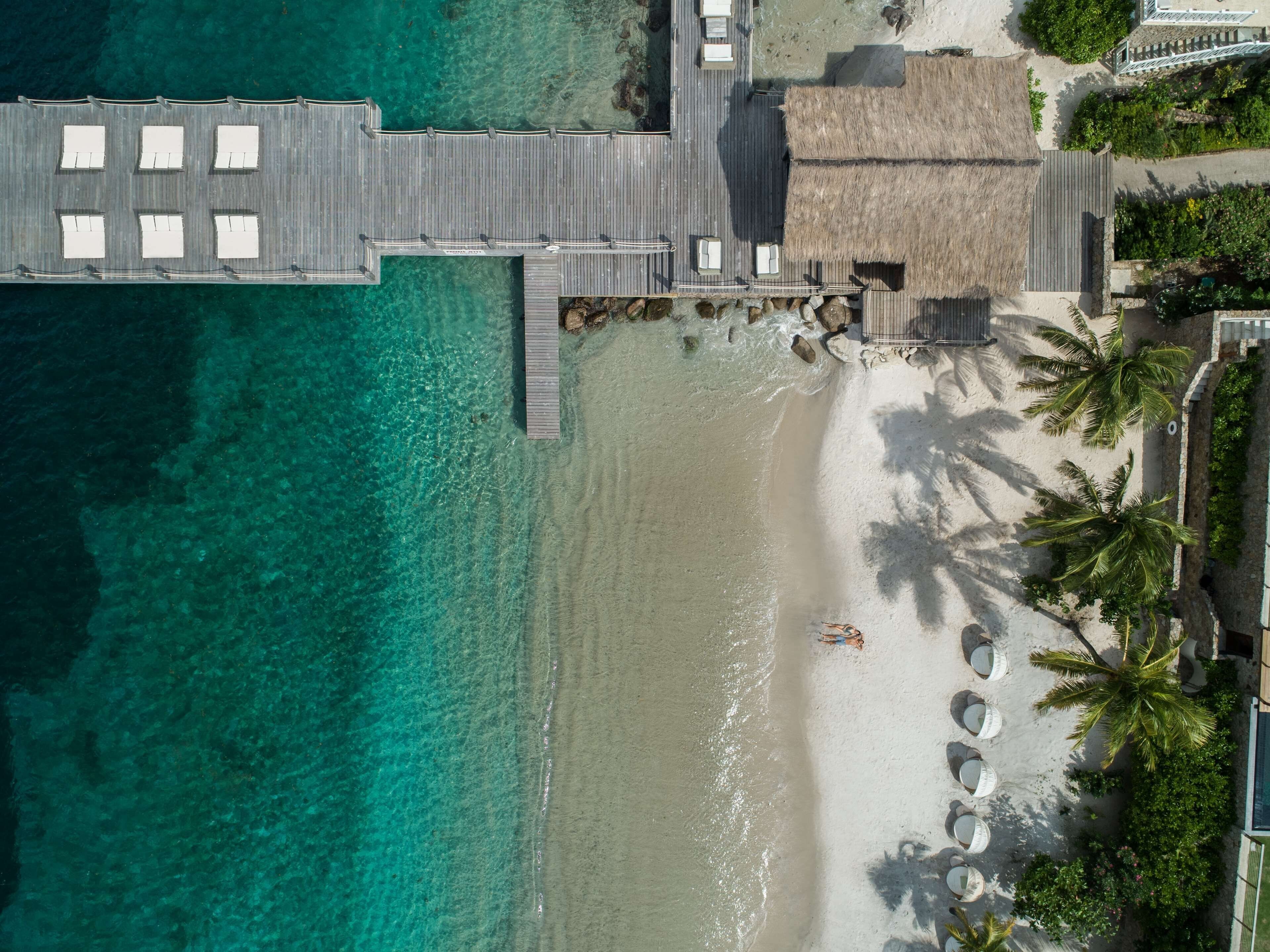 An aerial view of a beach resort with a wooden pier, beach umbrellas, and palm trees.