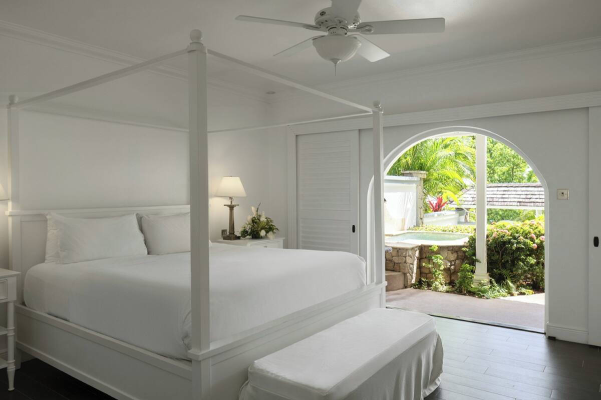 A four-poster bed with white bedding, two bedside tables with lamps, and a view of a garden through an arched doorway.
