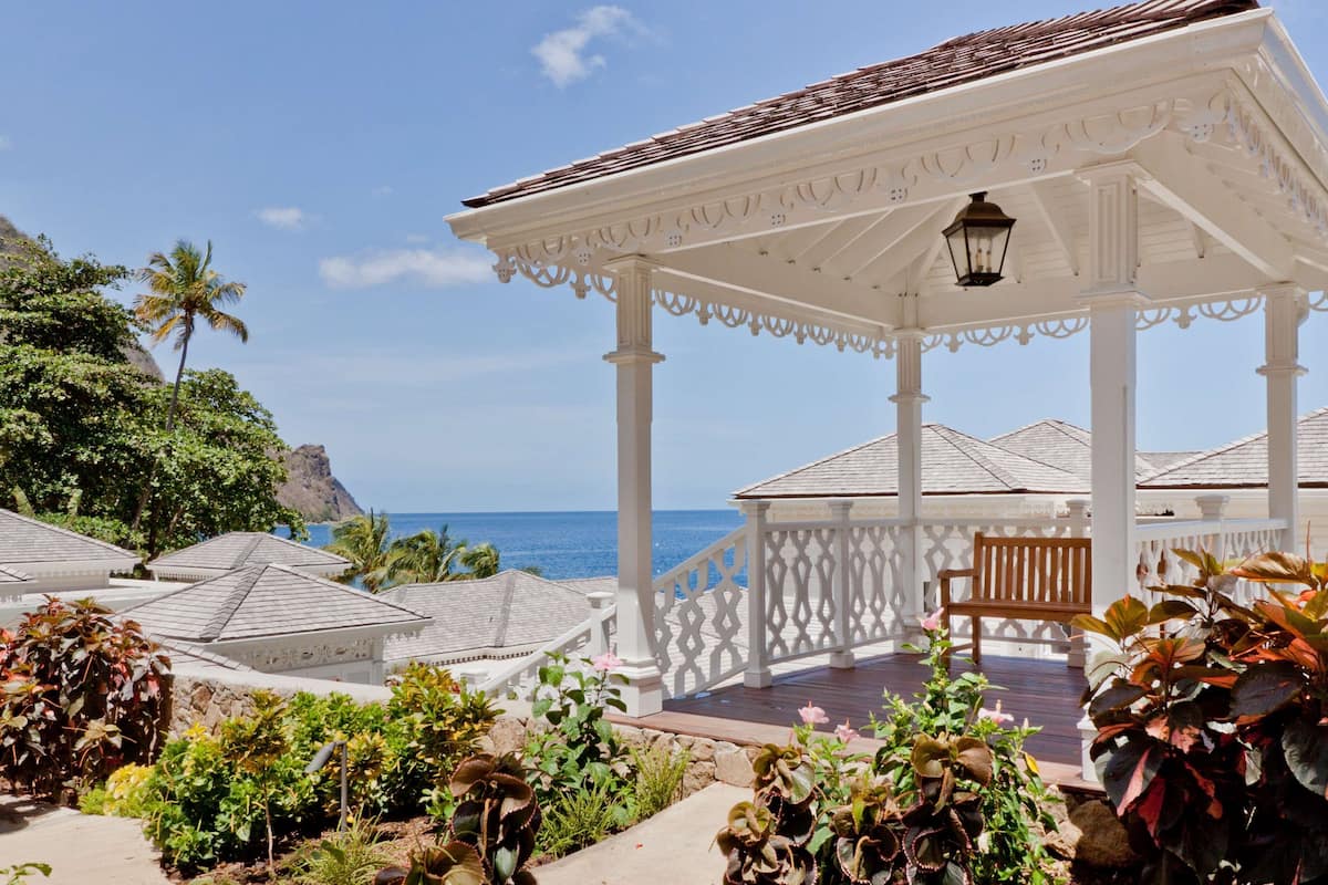 A covered outdoor seating area with a bench, surrounded by lush greenery and overlooking a coastal view.