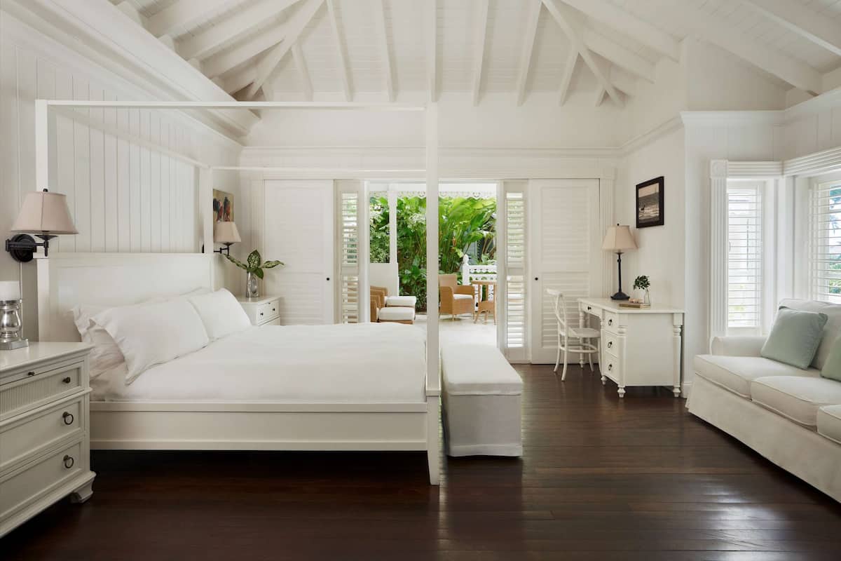 A white four-poster bed with a nightstand and lamp, a desk with a chair, and a sofa in a room with wooden floors and white walls.
