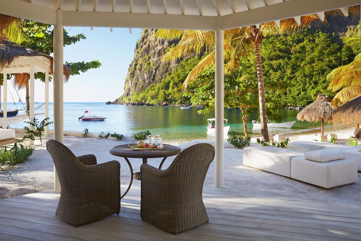 A covered outdoor seating area with wicker furniture, a round table, and a view of the sea and a mountain.