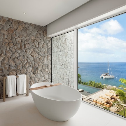 A modern bathroom with a stone wall, a freestanding bathtub, and a view of the ocean and boats.