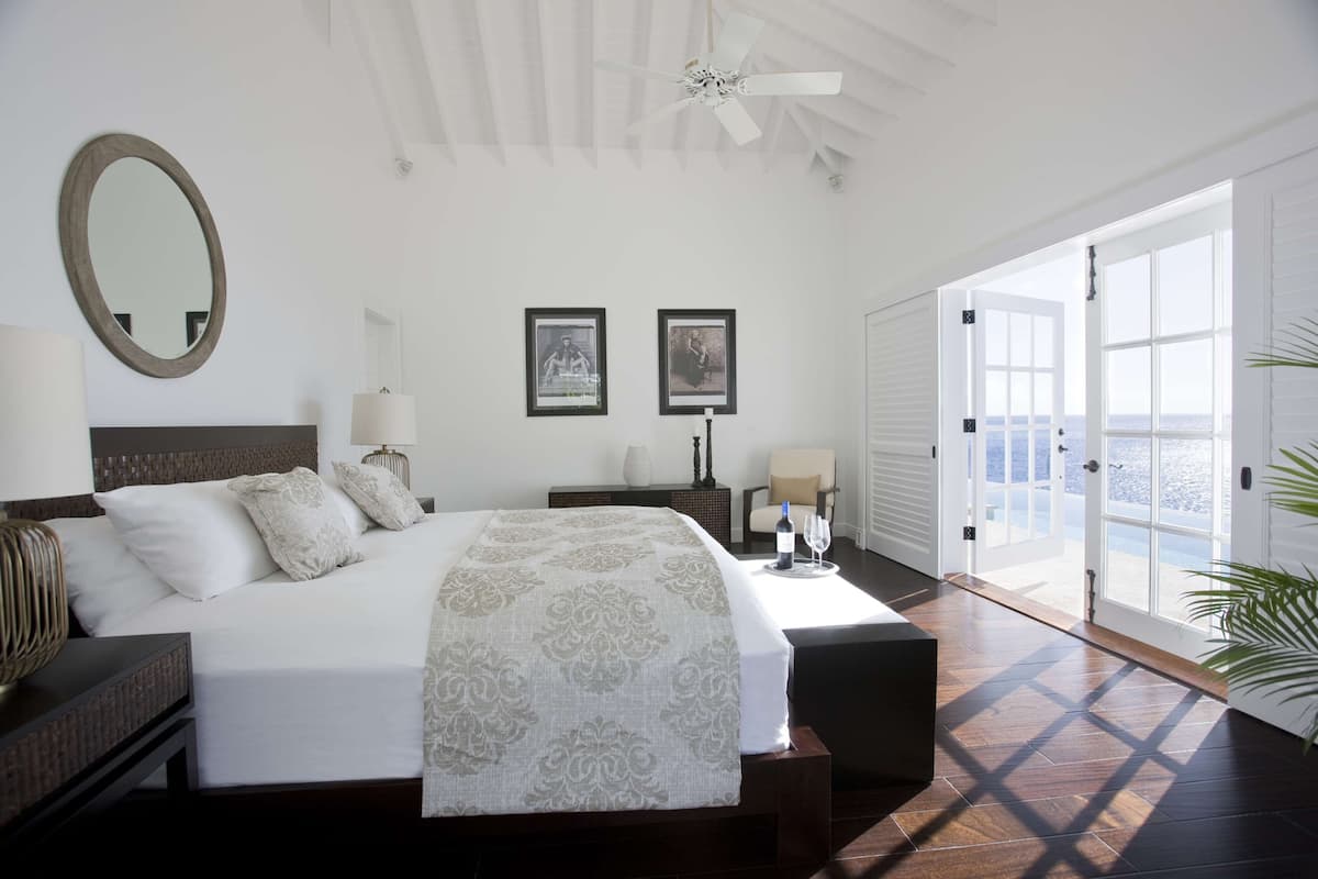 A bedroom with a large bed, bedside tables, a mirror, framed pictures, and a view of the sea through an open door.