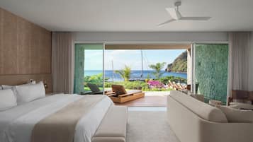 A modern hotel room with a large bed, a sofa, and a balcony with a view of the sea and boats.