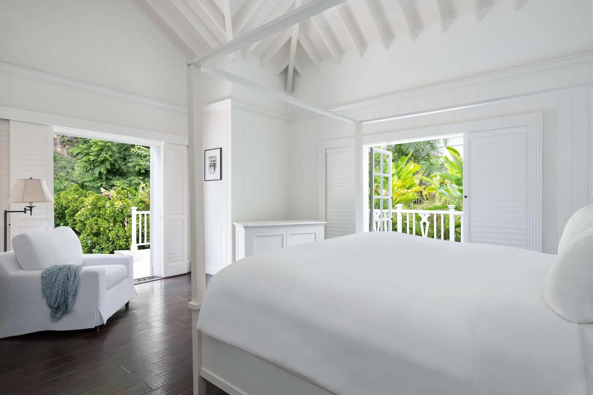 A white-themed bedroom with a large bed, a sitting area with a chair, and a view of lush greenery outside.