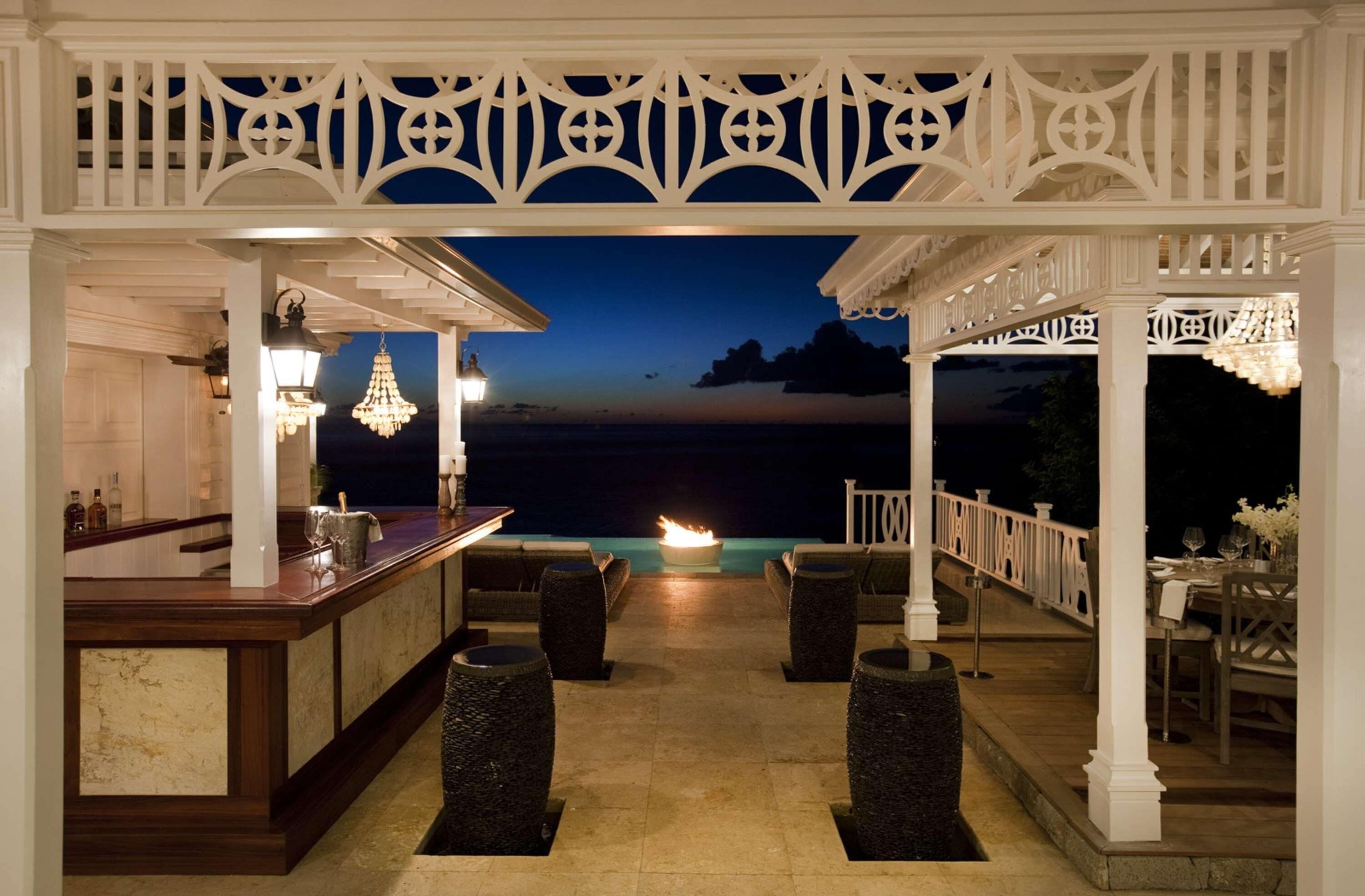 An outdoor bar area with a view of the ocean at dusk, featuring a white gazebo, wicker chairs, and a bar counter with bottles and glasses.