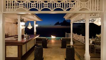 An outdoor bar area with a view of the ocean at dusk, featuring a white gazebo, wicker chairs, and a bar counter with bottles and glasses.