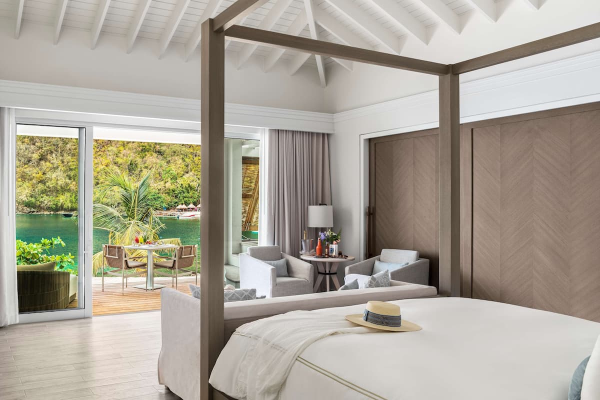 A spacious bedroom with a four-poster bed, a balcony with a view of a tropical garden and water body, and a seating area with a table and chairs.