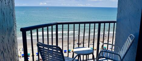 A balcony with two chairs and a small table, overlooking a beach with people and a parasail in the distance.