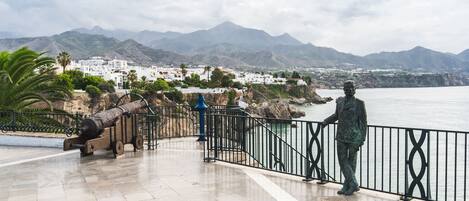 A coastal promenade with a statue, a cannon, and a view of a town and mountains.