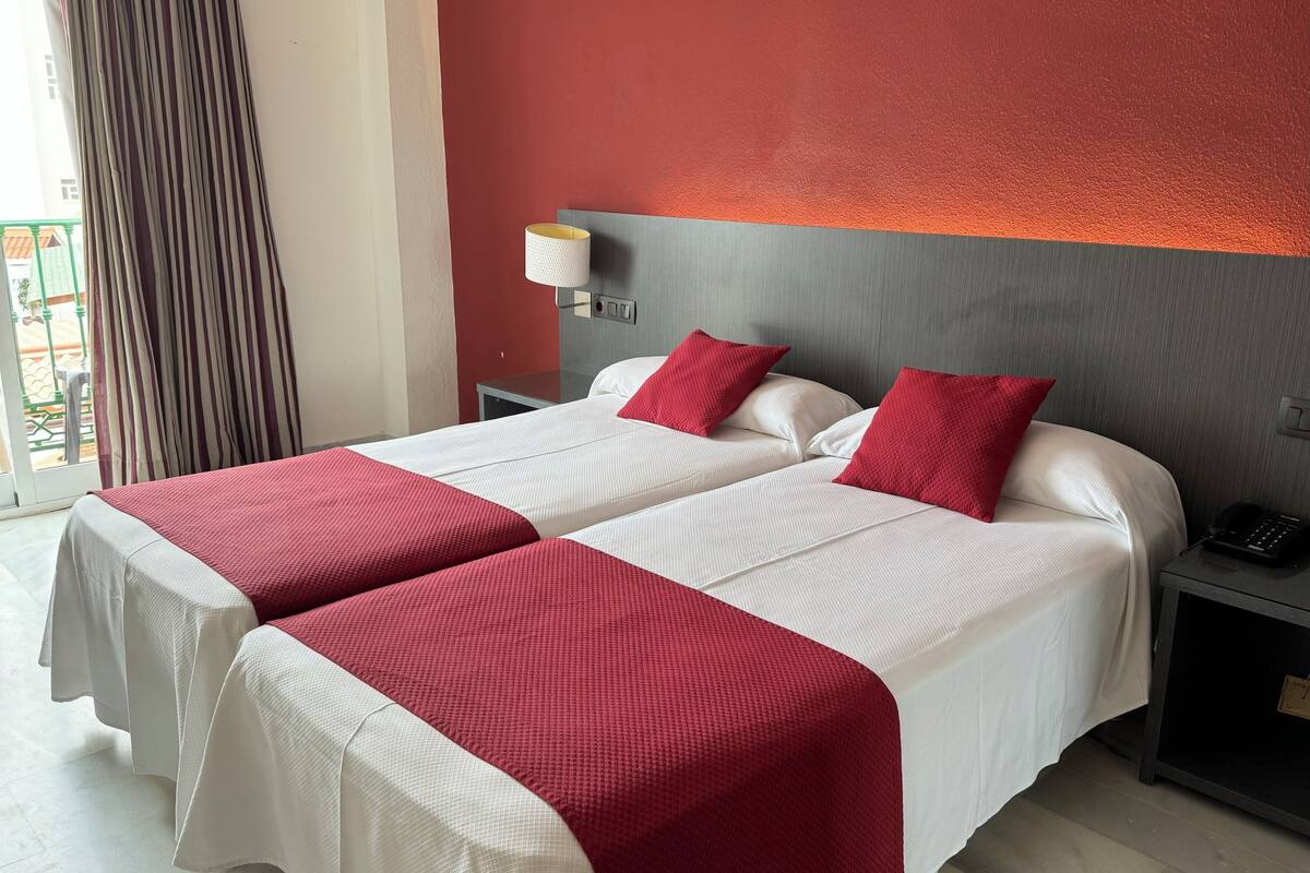 Habitación de hotel con dos camas, cada una con ropa blanca y detalles rojos, cabeceros grises, mesitas de noche con lámparas y vistas a un balcón.