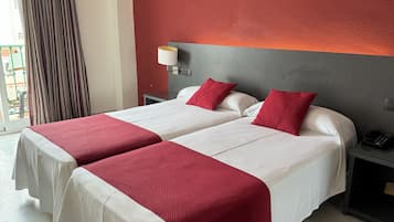 A hotel room with two beds, each with white bedding and red accents, a grey headboard, a bedside table with a lamp, and a view of a balcony.