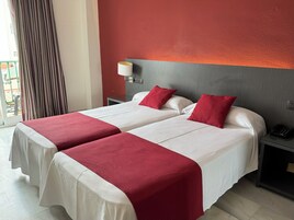 Habitación de hotel con dos camas, cada una con ropa blanca y detalles rojos, cabeceros grises, mesitas de noche con lámparas y vistas a un balcón.