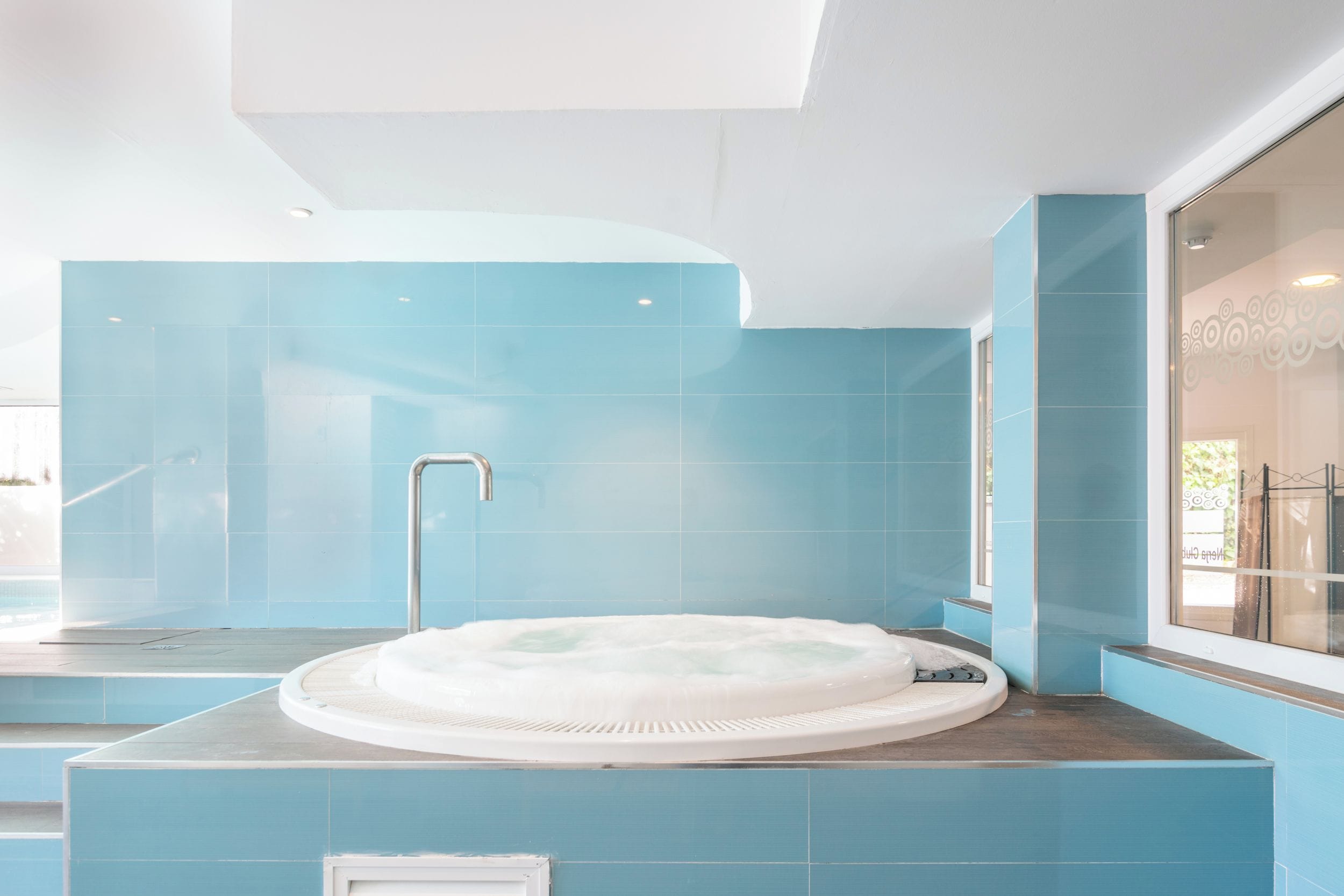 Een moderne badkamer met een centraal gelegen whirlpool, blauwe betegelde wanden en een groot raam.