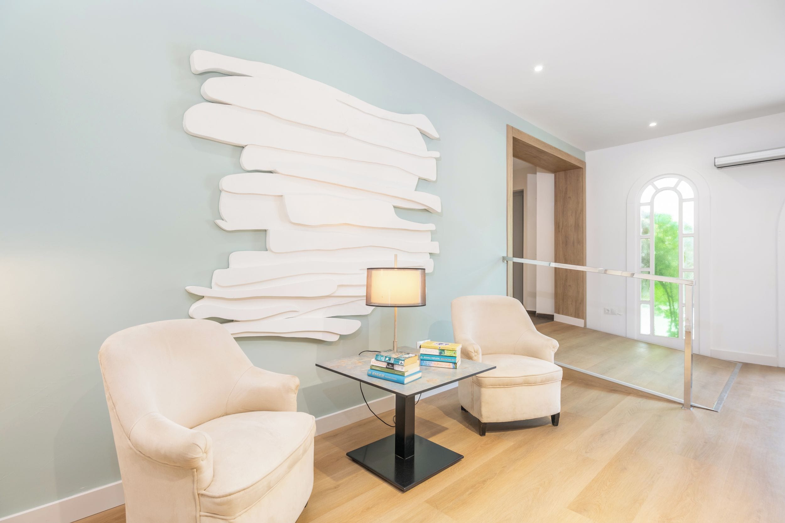 Una habitación moderna con pared azul claro, una mesa de vidrio con libros, dos sillones blancos y un ventanal grande.