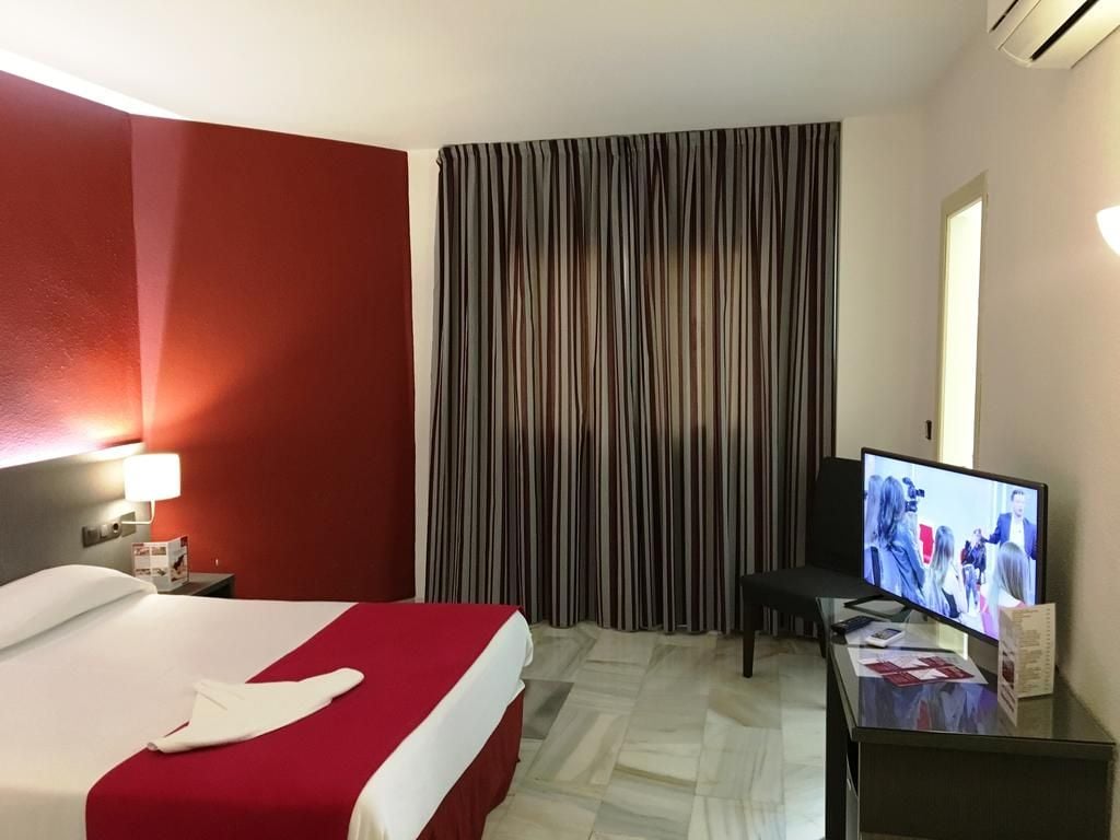 A hotel room with a bed, a desk with a television, a chair, and a window with curtains.