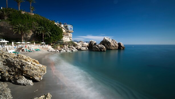 A coastal beach with a rocky shoreline, palm trees, and a building with a balcony overlooking the sea. - Nerja Club by Dorobe (Nerja)
