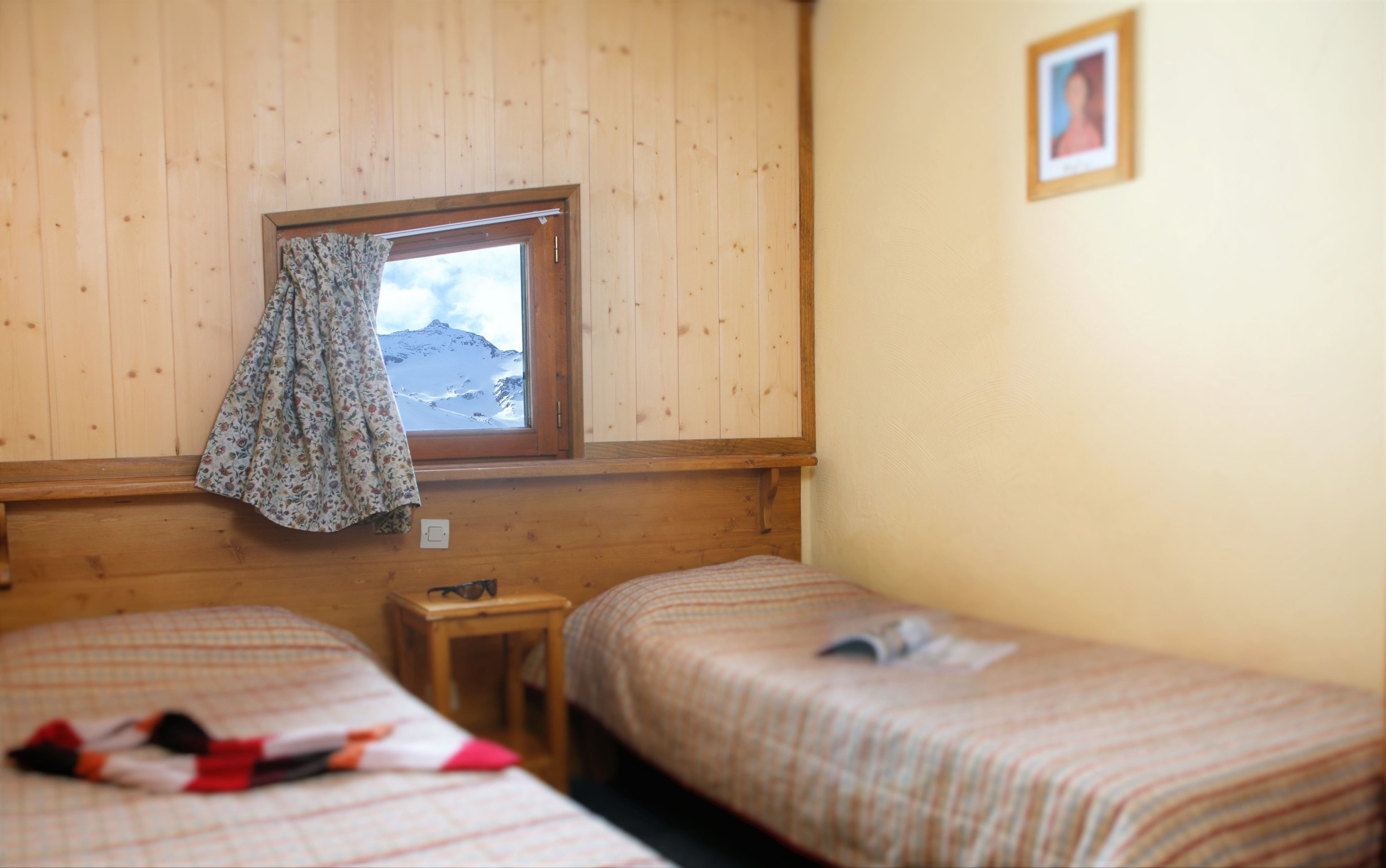 Ein Zimmer mit zwei Betten, einem Fenster mit Vorhang und einem gerahmten Bild an der Wand.